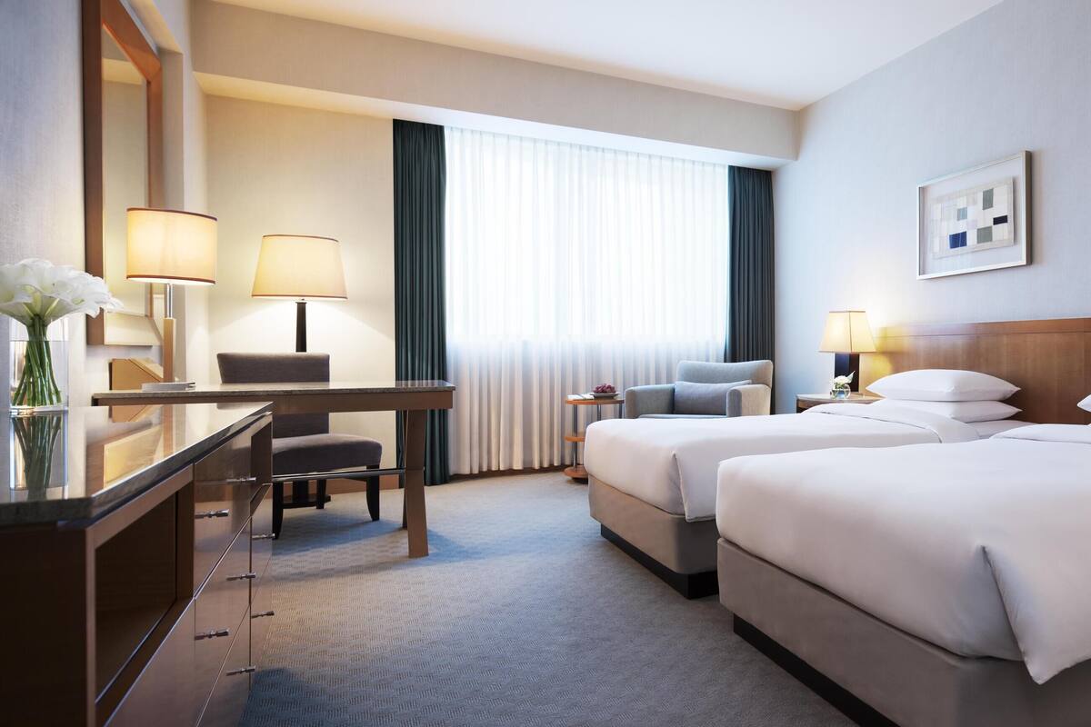 Une chambre d’hôtel avec deux lits, un bureau, une chaise, une commode et une grande fenêtre avec des rideaux.