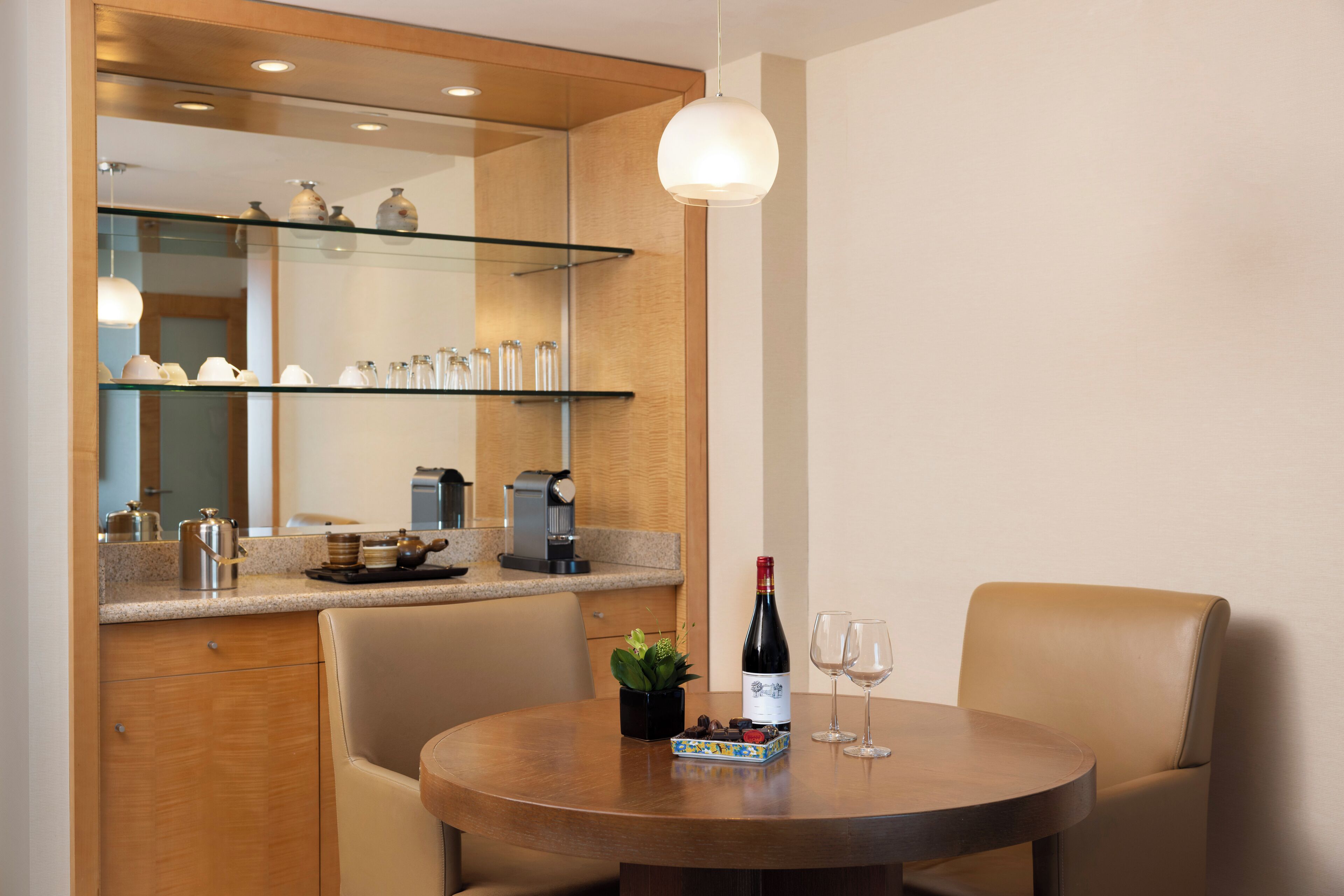 A modern dining area with a round table, two chairs, a bottle of wine, and two glasses. A small plant and a decorative box are on the table. A glass shelf above the table holds various items.