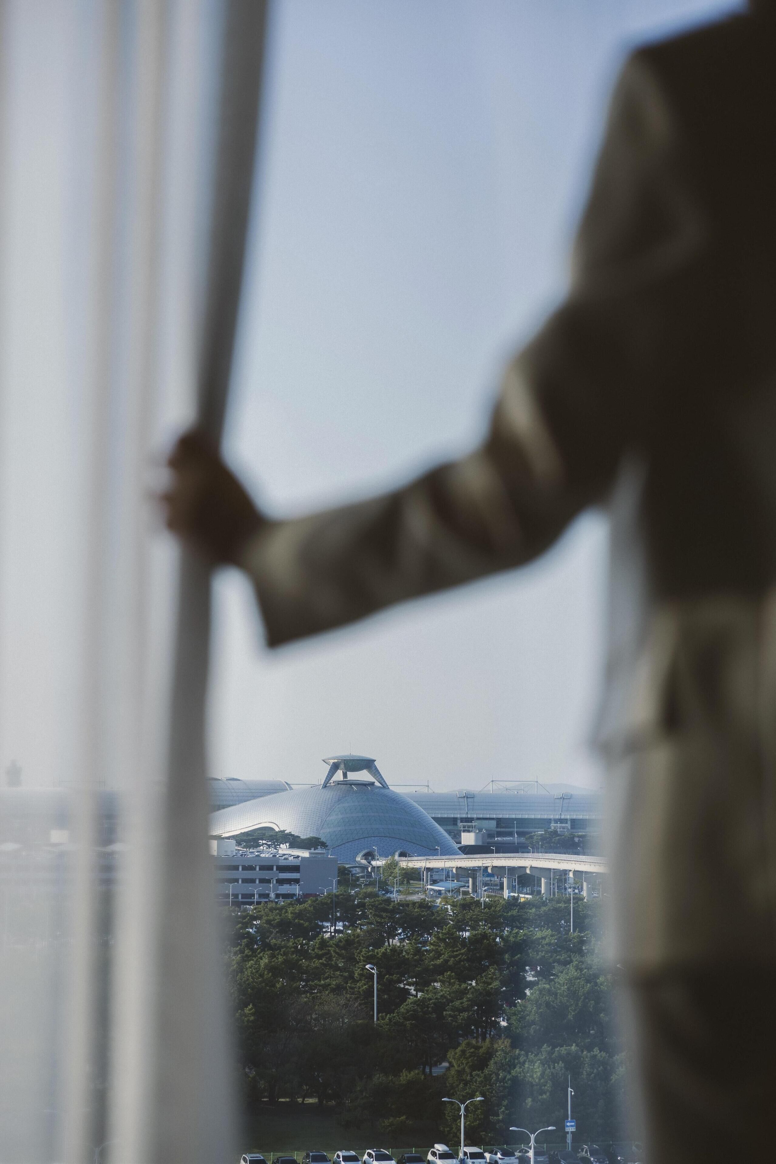 Una persona vestida con traje, de pie junto a una ventana con vista a un edificio moderno y un puente.
