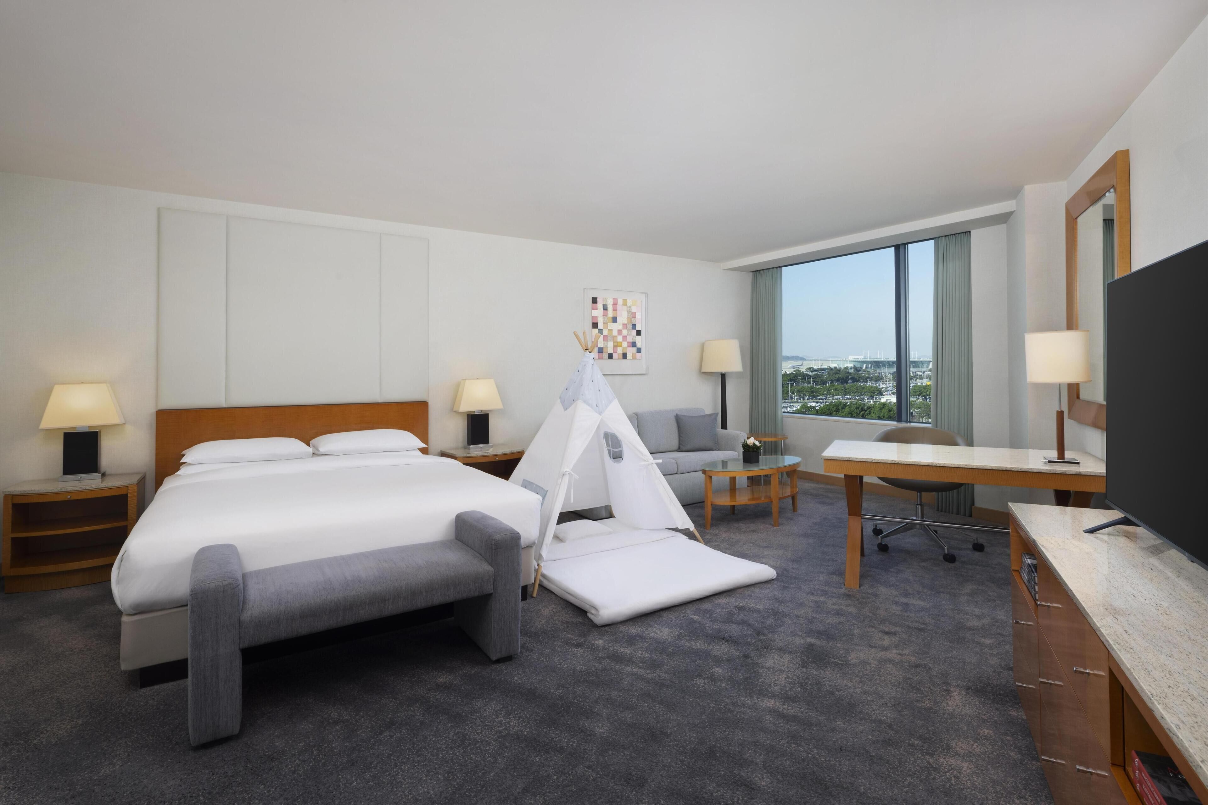 A hotel room with a bed, a desk, a chair, a TV, a lamp, a window, and a painting.