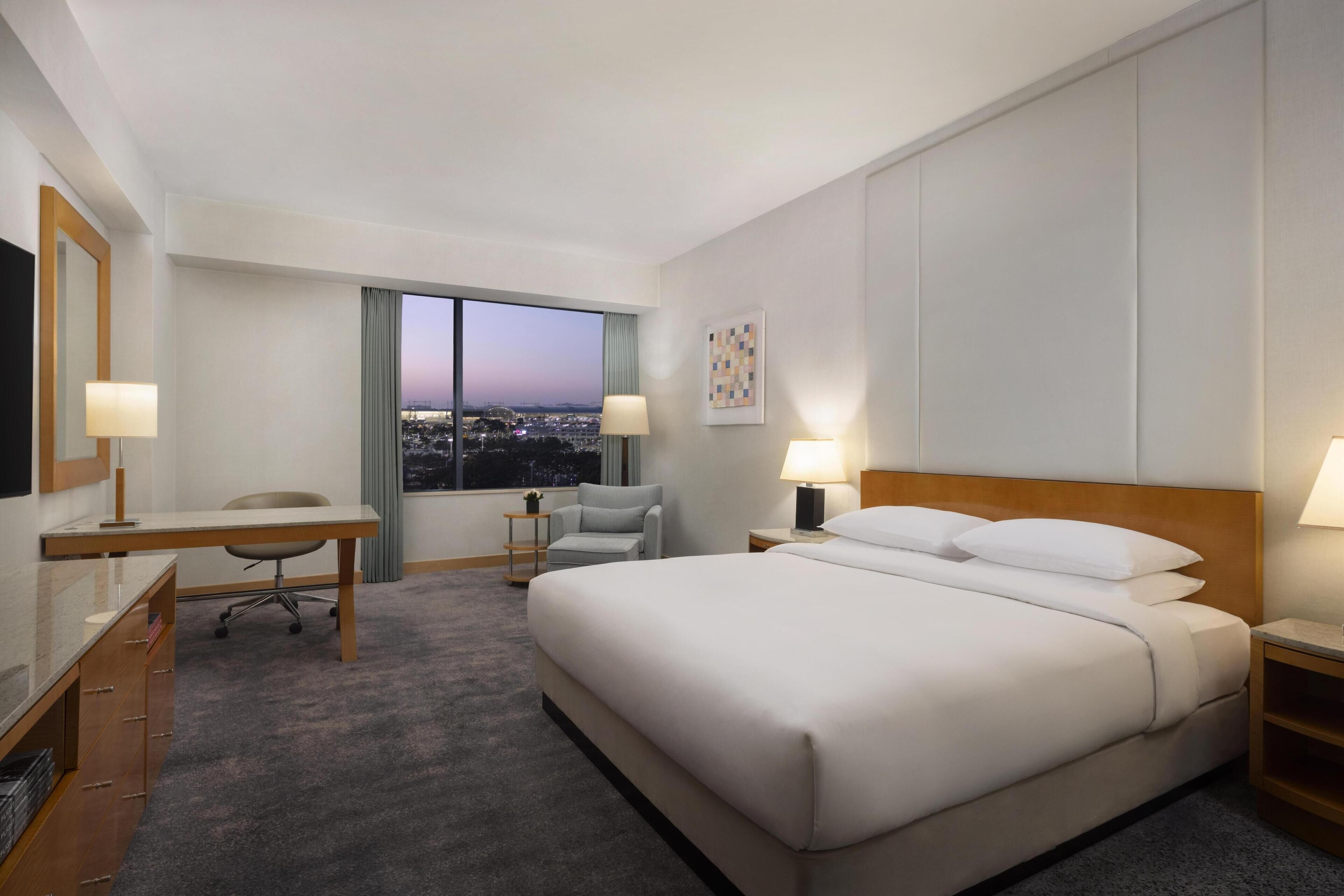 A hotel room with a large bed, a desk, a chair, a small table, and a window with a city view.