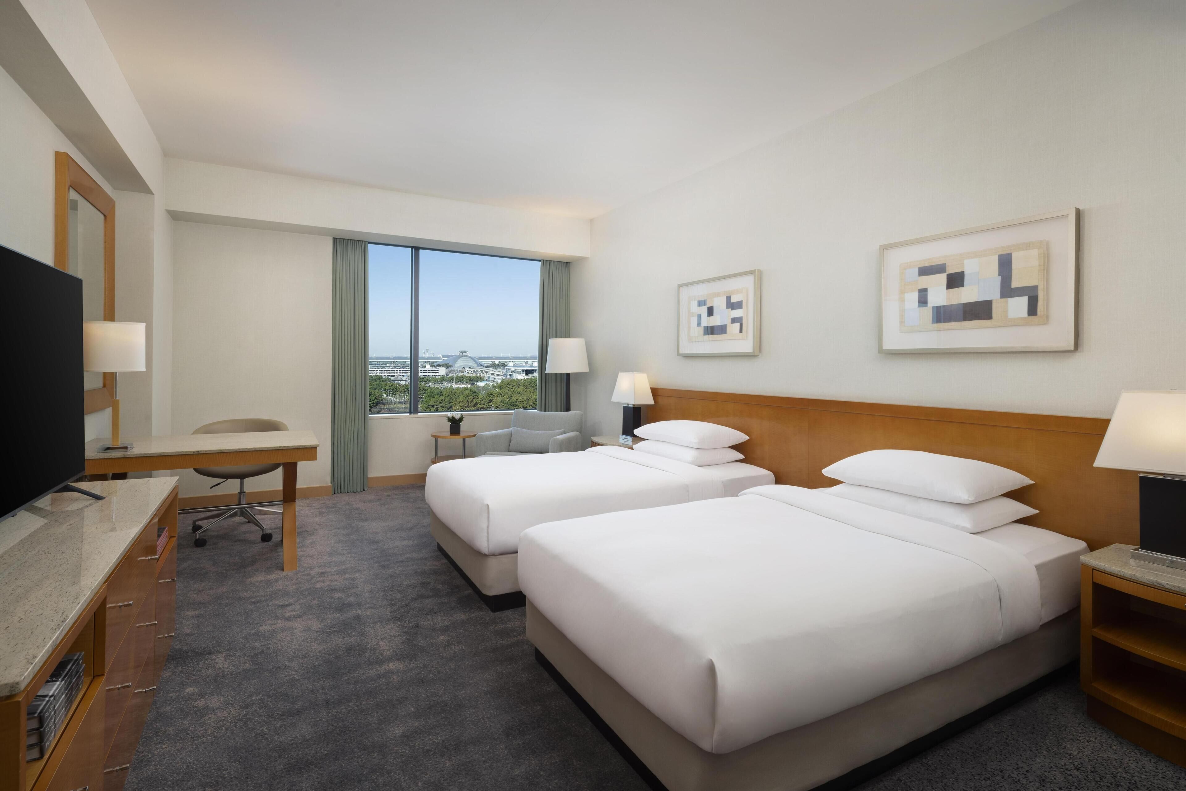 Quarto de hotel com duas camas, uma escrivaninha, uma TV e uma janela grande com vista para a cidade.