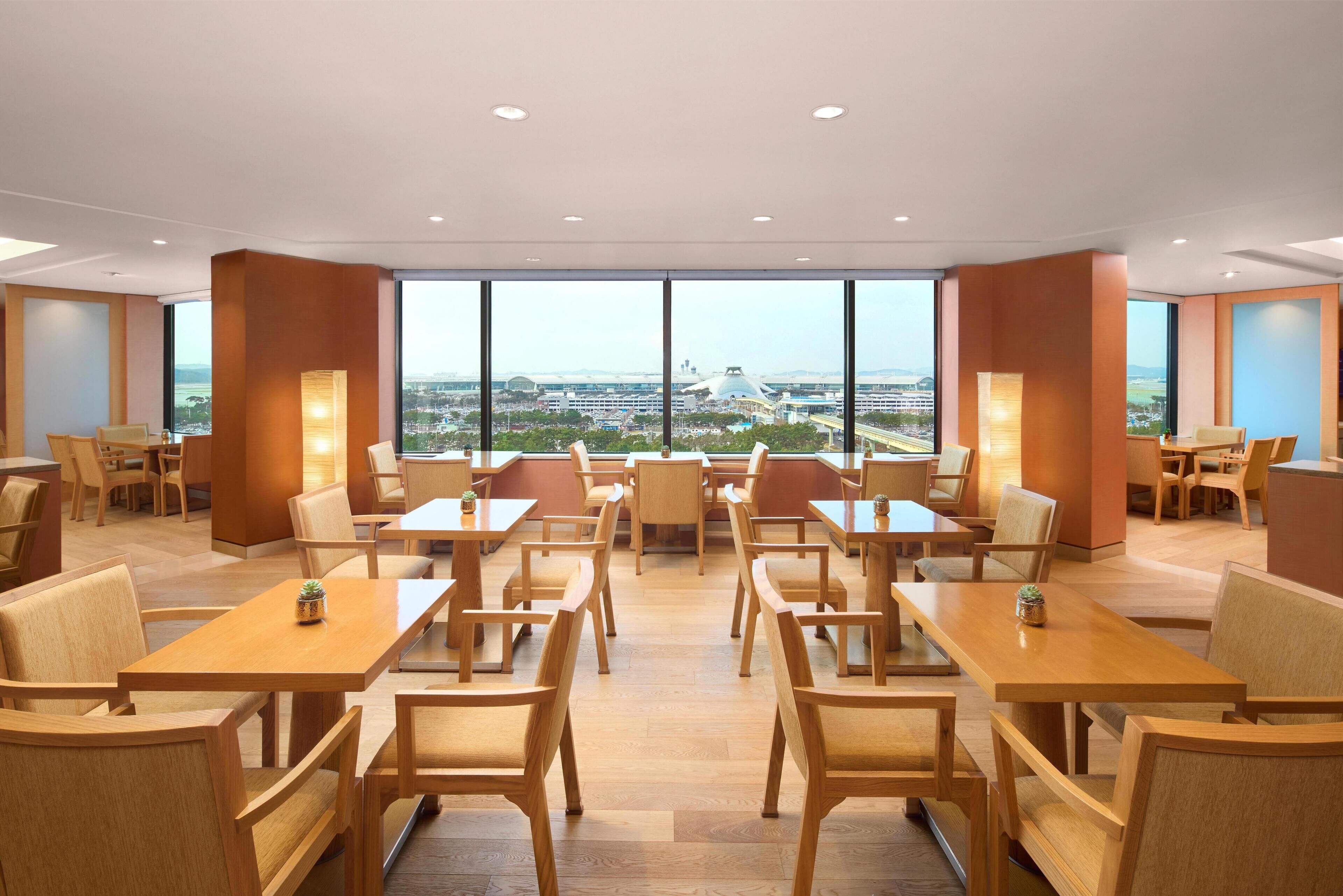 Une salle à manger d’hôtel avec de nombreuses tables et chaises, et de grandes fenêtres donnant sur la ville.