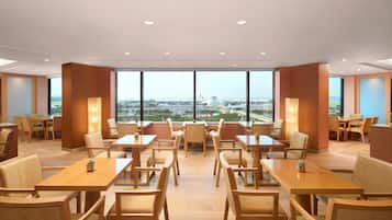 Une salle à manger d’hôtel avec de nombreuses tables et chaises, et de grandes fenêtres donnant sur la ville.