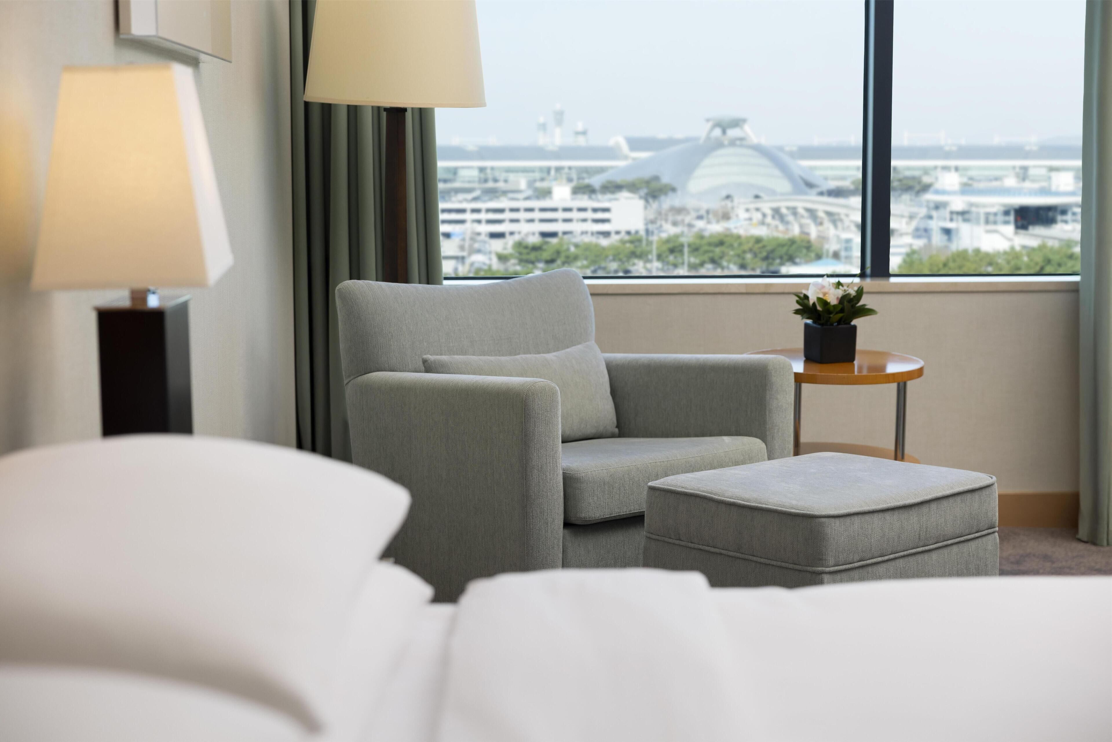 A hotel room with a grey sofa, a small ottoman, a side table with a plant, and a view of a cityscape through large windows.