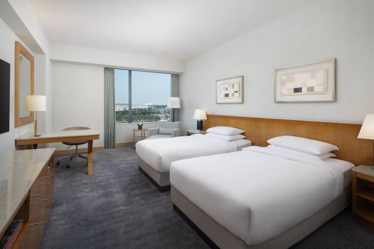Une chambre d’hôtel avec deux lits, un bureau, une chaise, une télévision et une grande fenêtre donnant sur la ville.