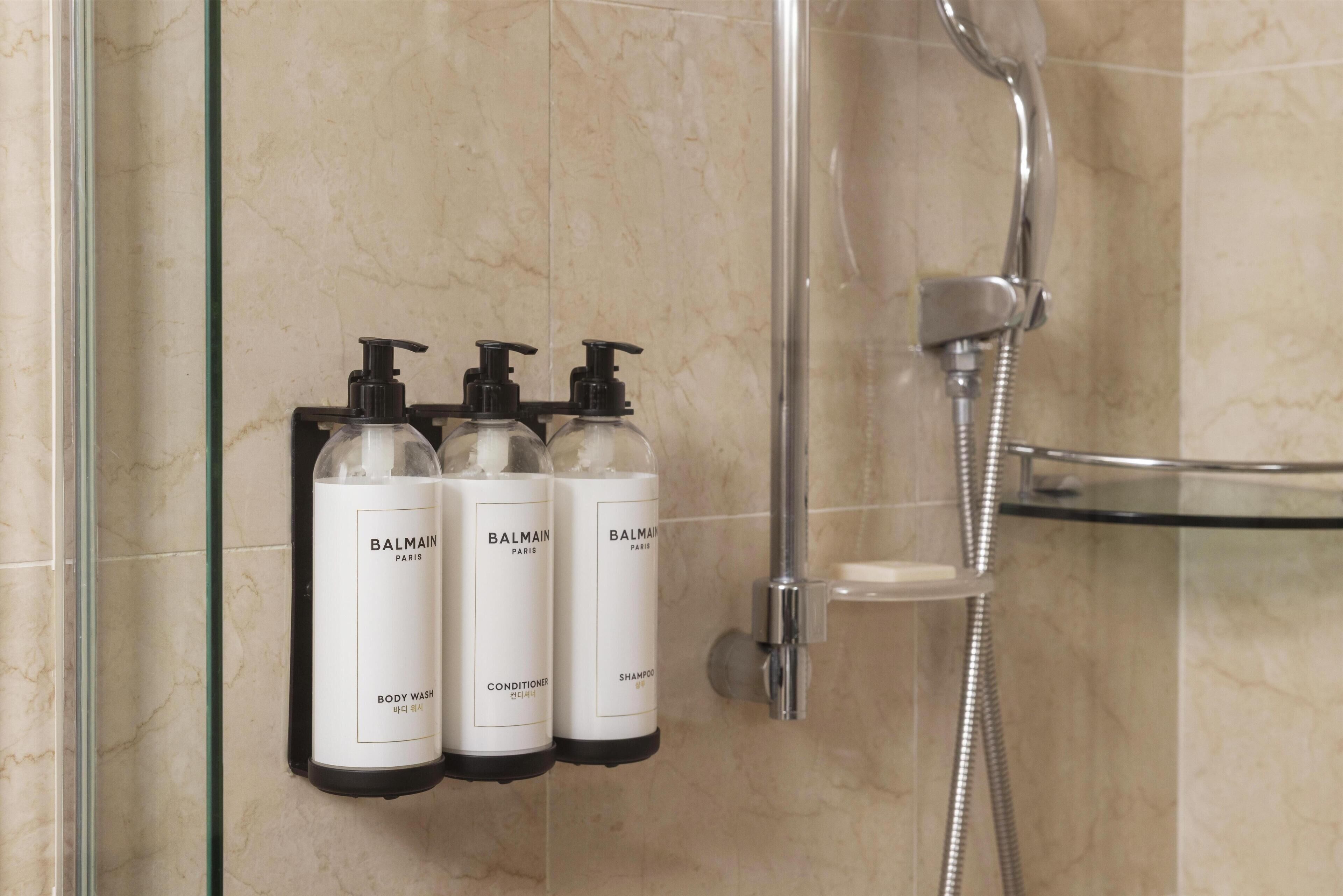 A bathroom with a glass shower enclosure, a handheld showerhead, and three Balmain Paris toiletry bottles mounted on the wall.