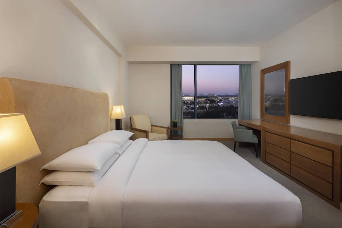 Une chambre d’hôtel équipée d’un lit, de lampes de chevet, d’un bureau, d’une chaise, d’une télévision et d’une grande fenêtre donnant sur la ville.