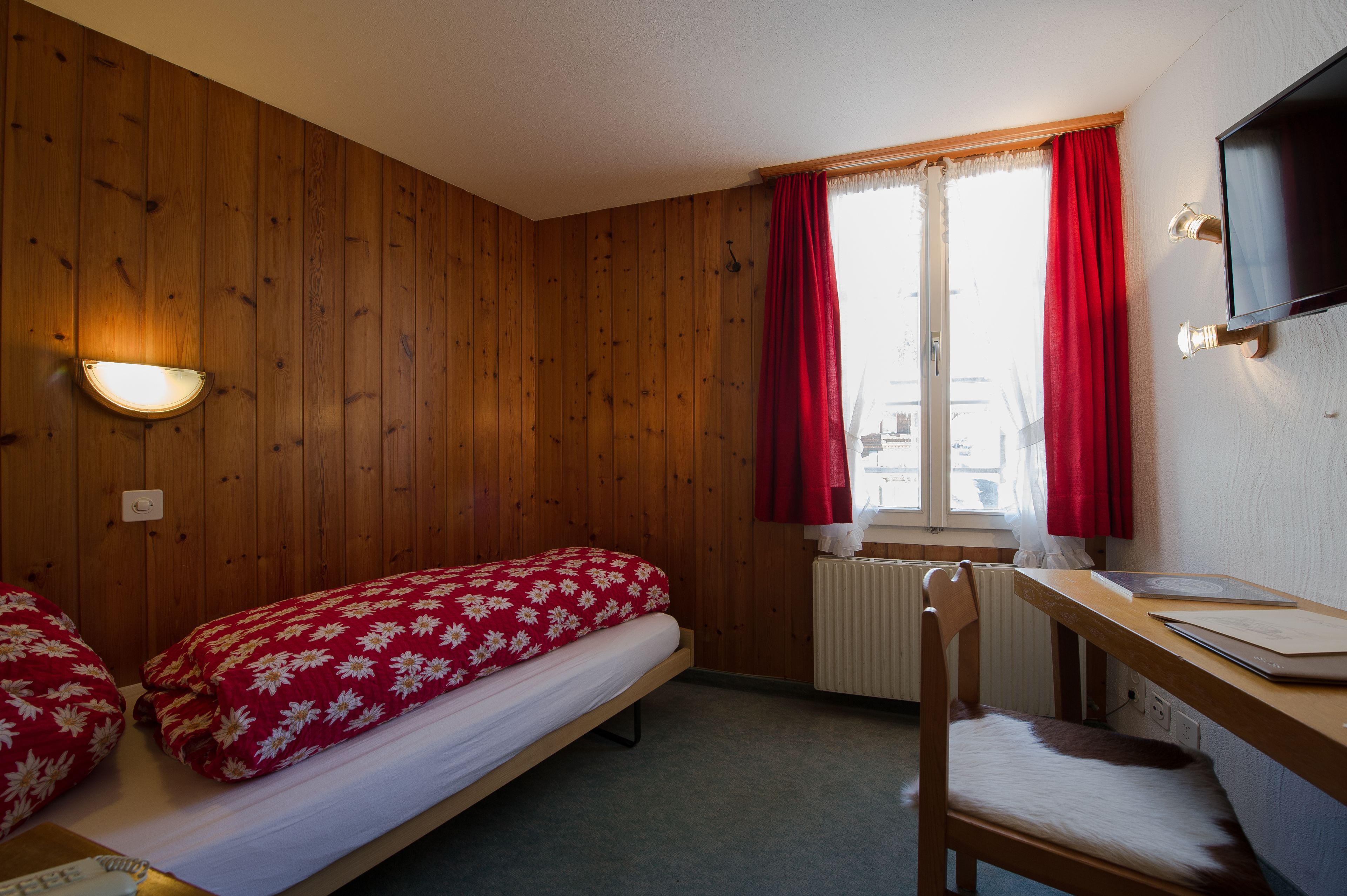 A room with wooden walls, a bed with a red floral bedspread, a desk with a chair, a window with red curtains, and a mounted TV.