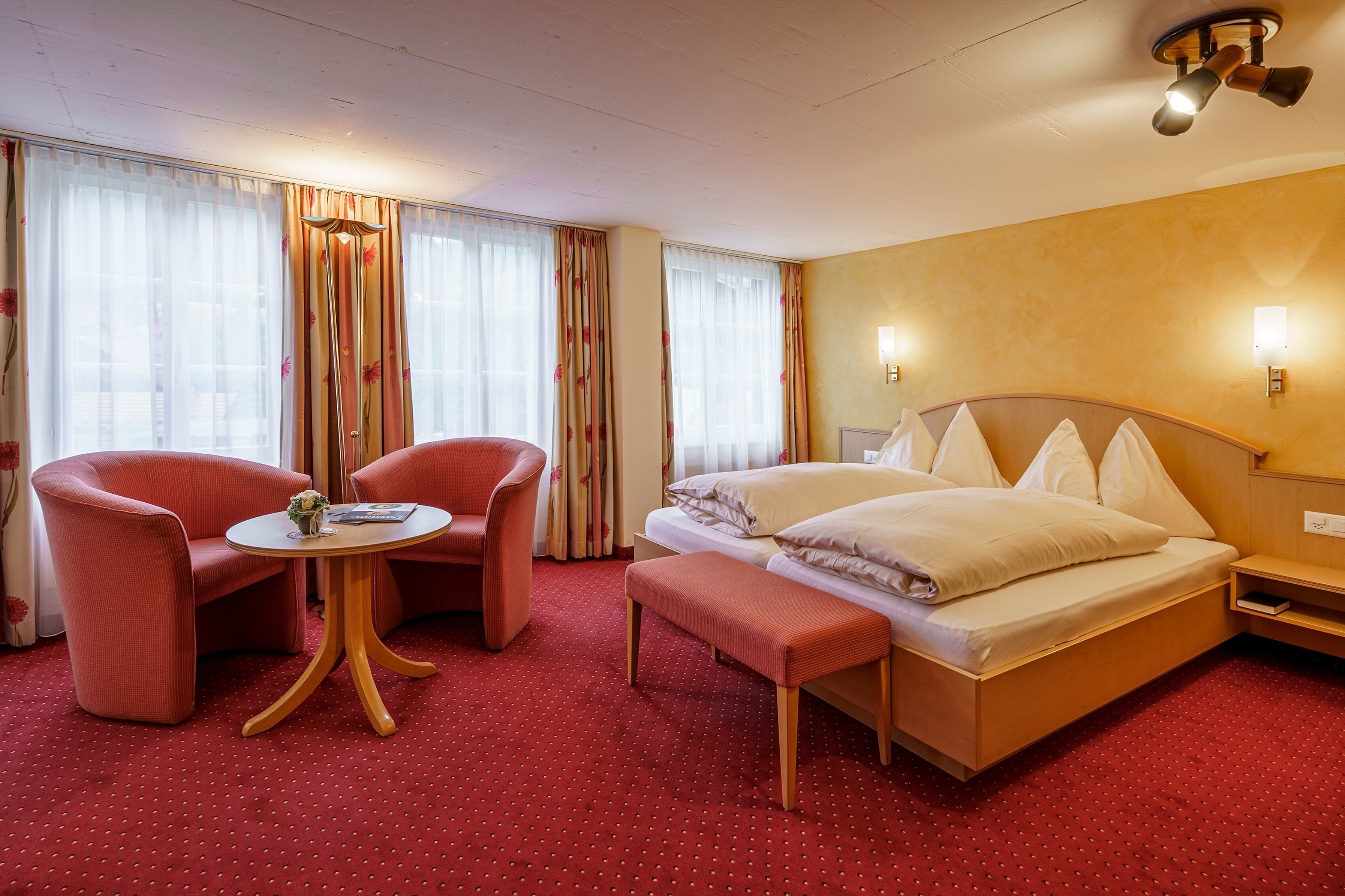 A hotel room with a bed, two red armchairs, a small round table, and a bench.