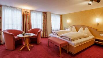 A hotel room with a bed, two red armchairs, a small round table, and a bench.