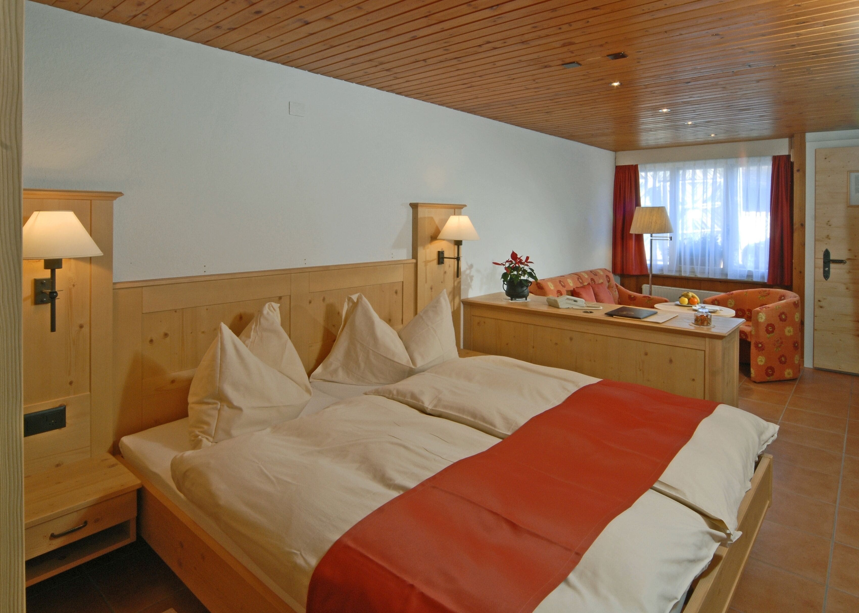 A hotel room with a large bed, wooden headboard, two bedside tables with lamps, a desk with a chair, and a window with curtains.