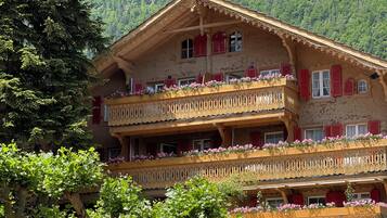 Un hôtel de style alpin traditionnel, doté de balcons ornés de jardinières, d’un mur en pierre agrémenté d’une étagère à vin et offrant une vue dégagée sur les collines boisées environnantes.