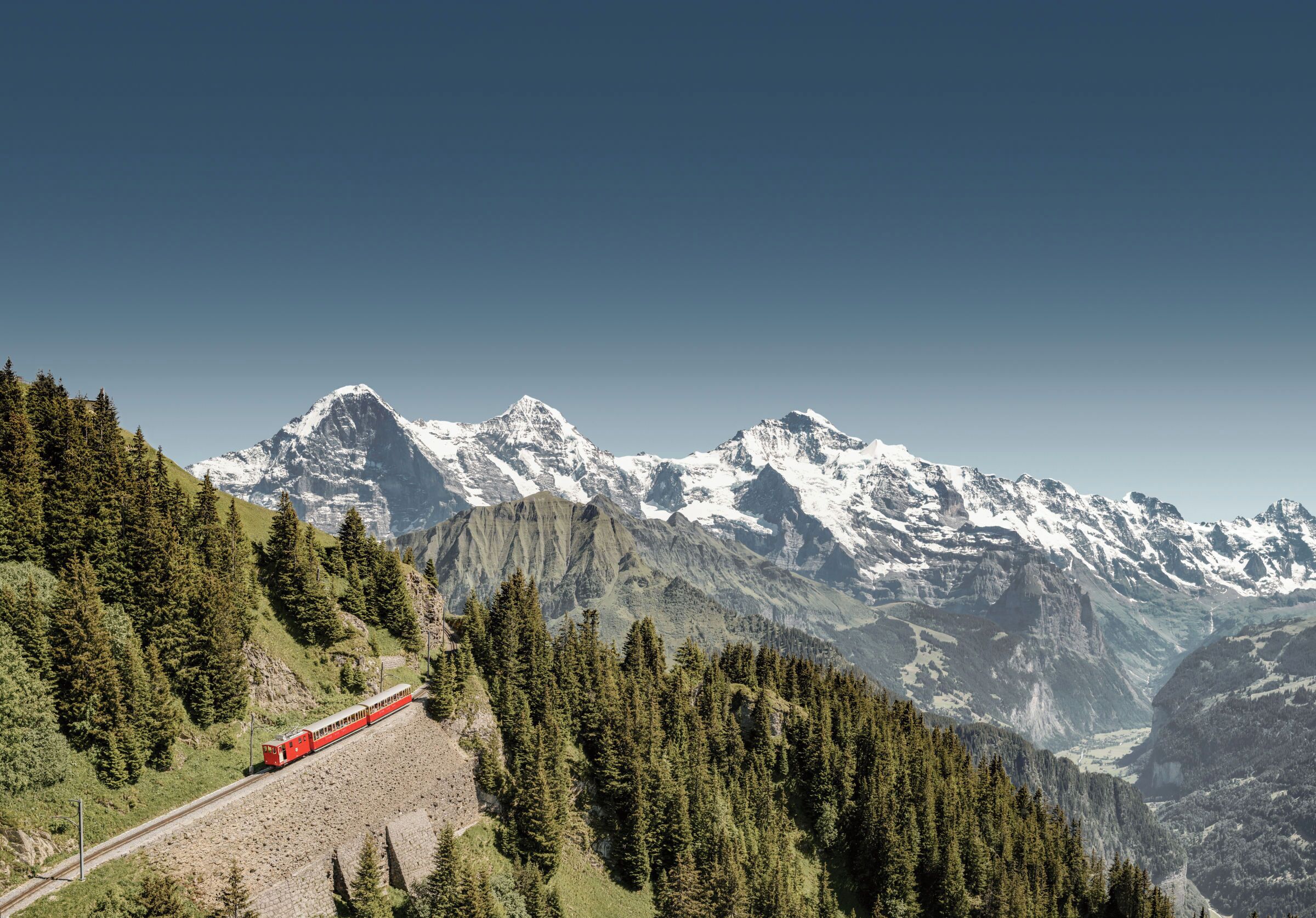 A red train traveling through a mountainous area with snow-capped peaks and dense forests.
