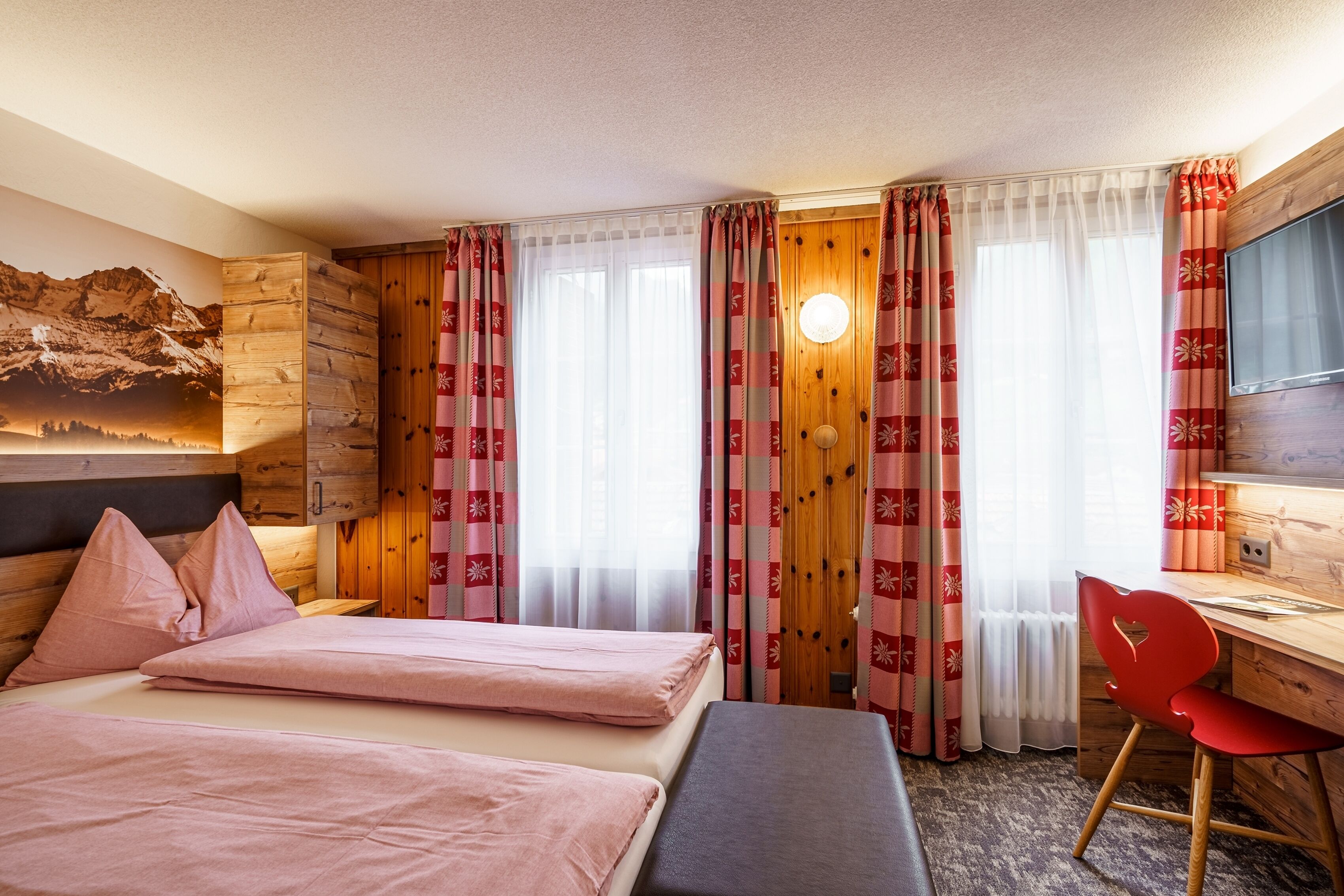 Habitación de hotel con cama, escritorio y silla. Amplia ventana con cortinas, televisor montado en la pared y cabecero de madera.