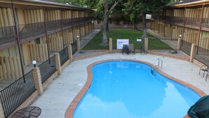 Una piscina con borde de ladrillo, rodeada por una cerca y un patio cubierto con sillas y una mesa.