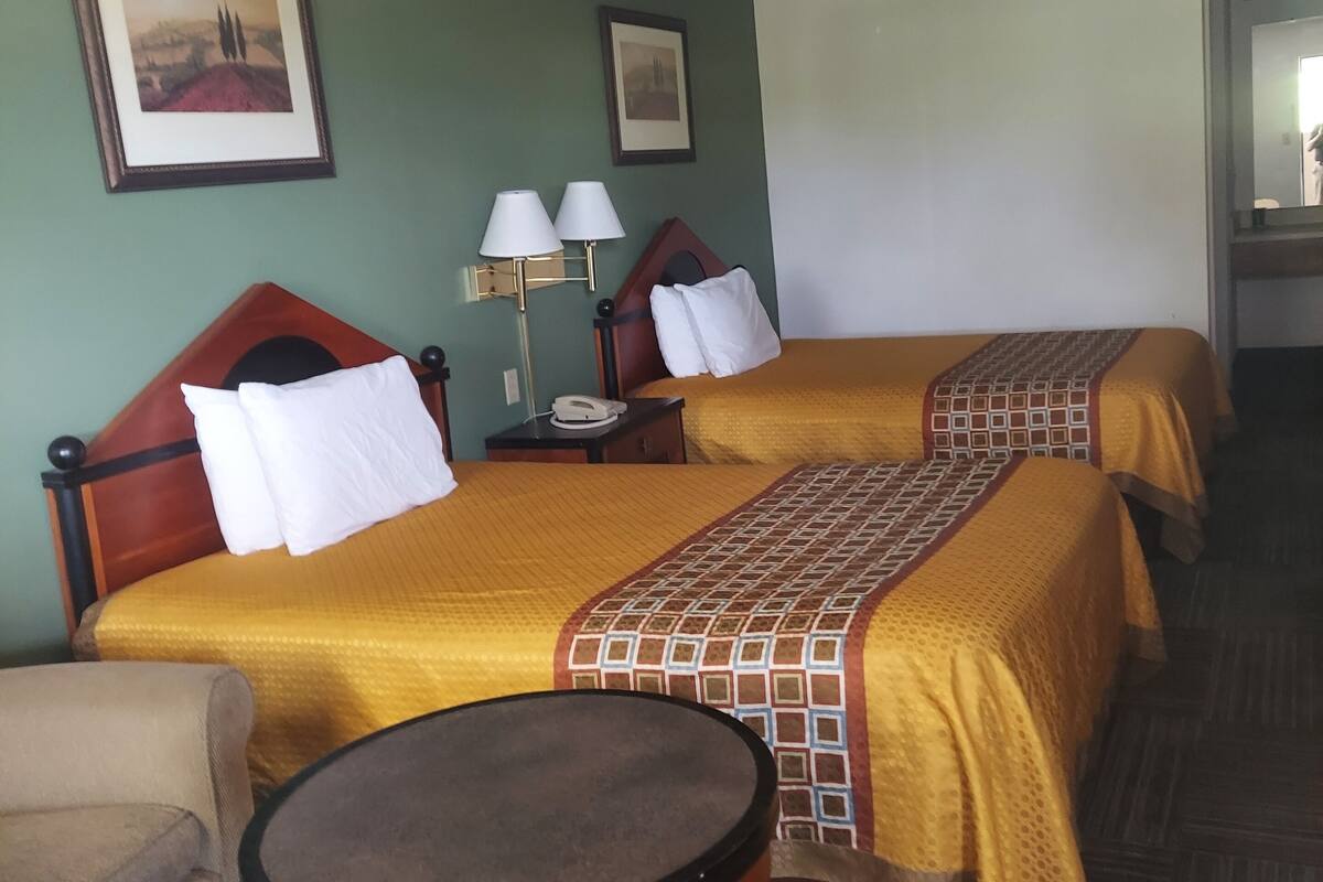 A hotel room with two beds, a round table, a chair, and a lamp.
