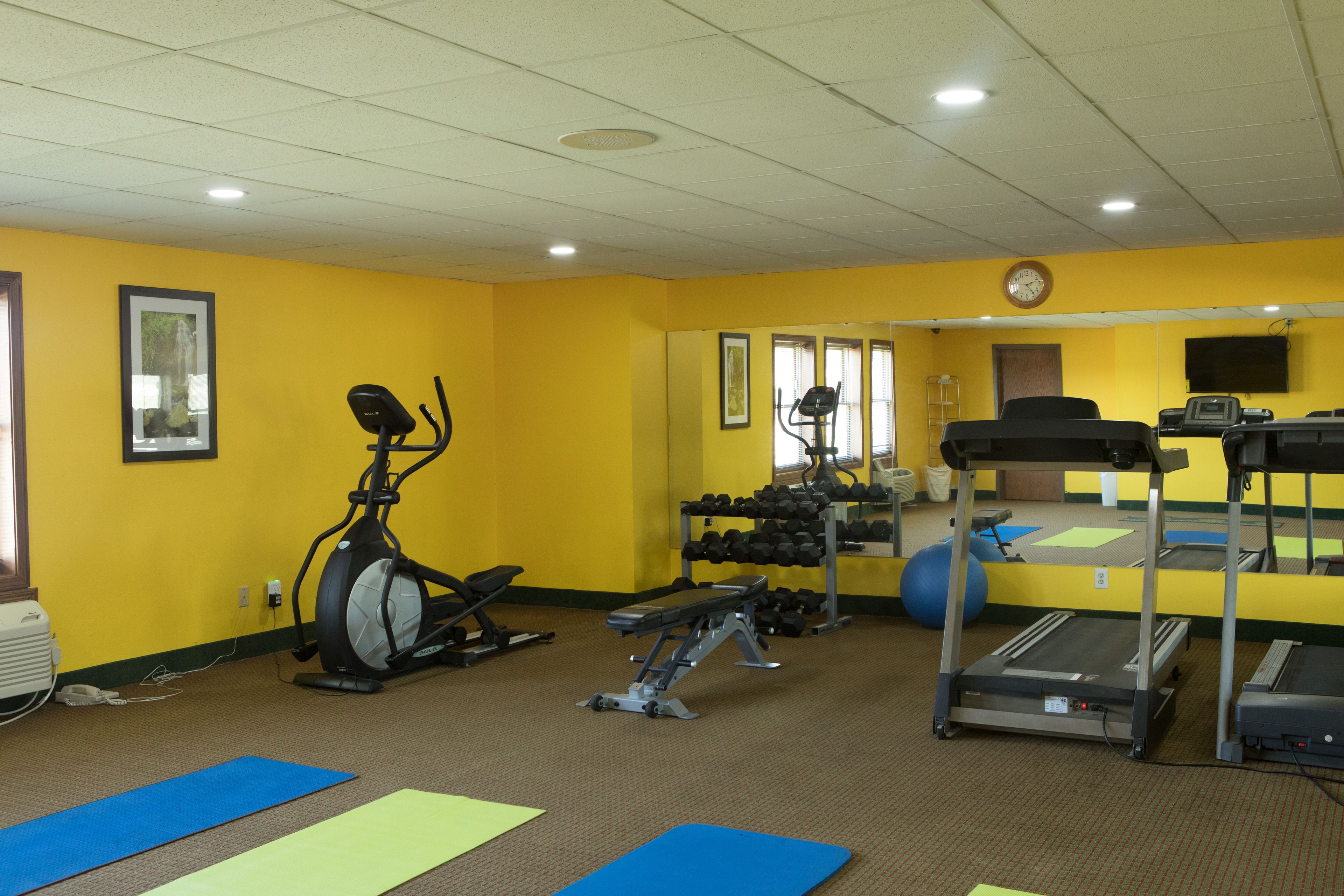 Academia com equipamentos de ginástica, tapetes e um relógio na parede.