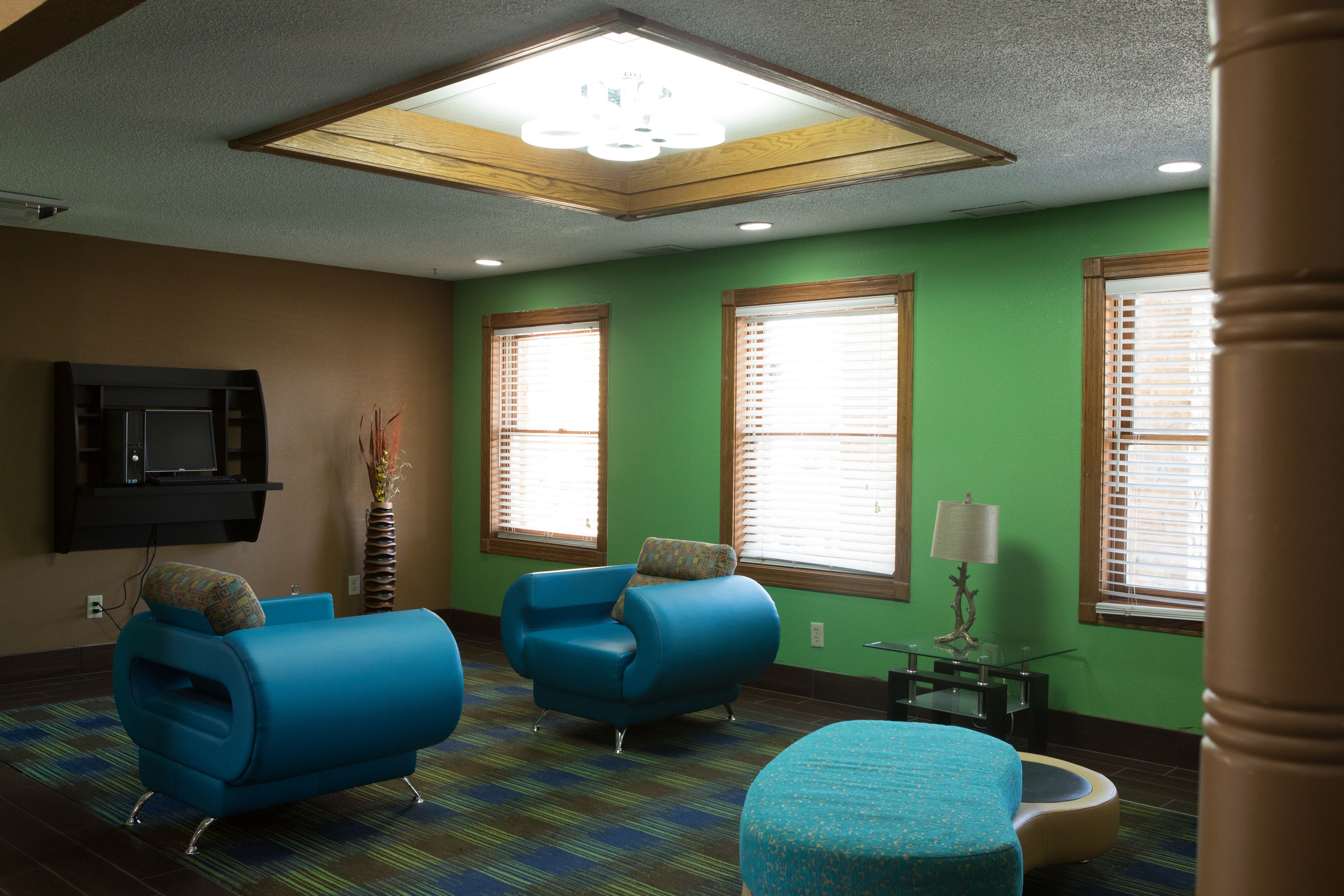 A room with two blue armchairs, a small ottoman, a television mounted on the wall, and a window with blinds.