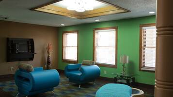 A room with two blue armchairs, a small ottoman, a television mounted on the wall, and a window with blinds.