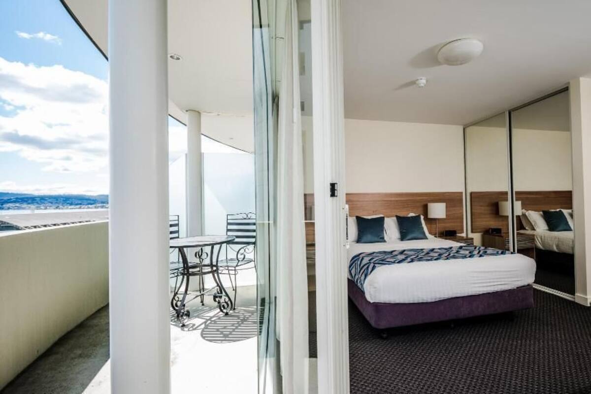 A hotel room with a bed, a small table, a chair, a bench, and a large mirror.