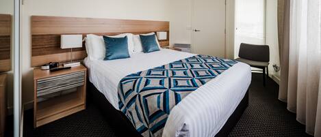 A hotel room with a large bed, a wooden headboard, a desk with a lamp, a chair, and a TV.