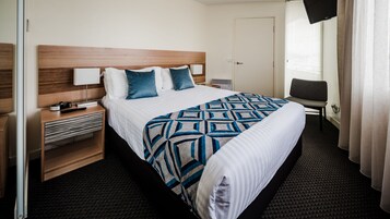 Una habitación de hotel con una cama grande, cabecera de madera, un escritorio con lámpara, una silla y un televisor.