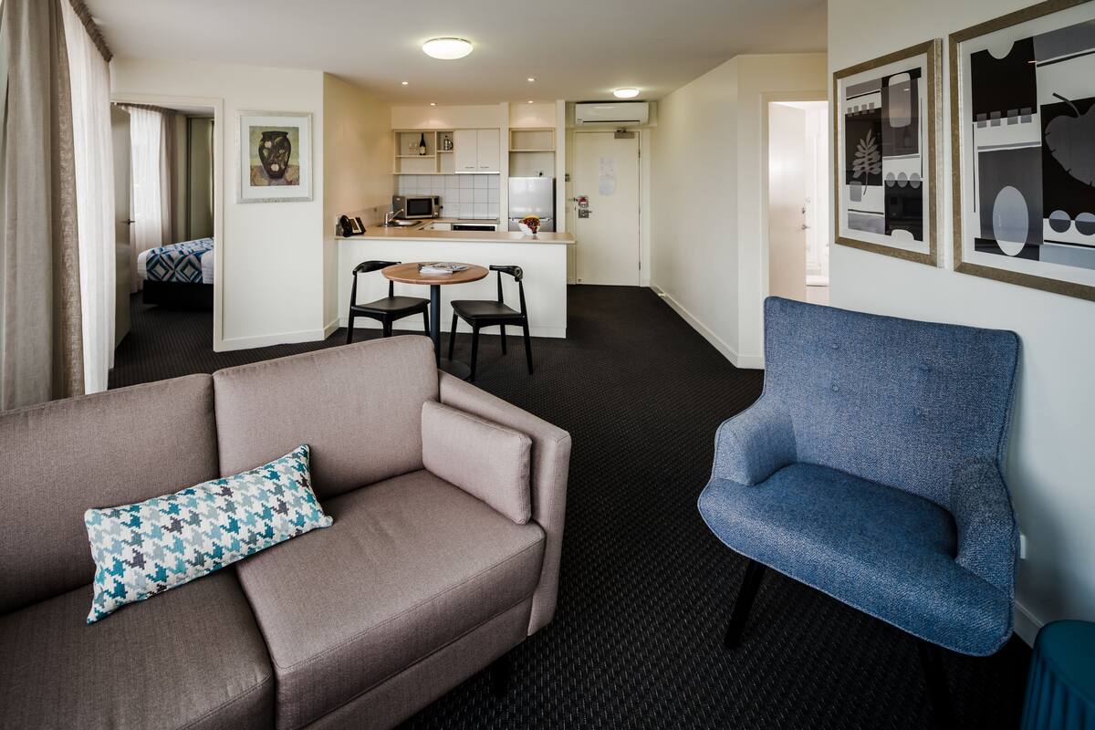 A modern hotel room with a sofa, a blue armchair, a dining area with a table and chairs, and a kitchenette.
