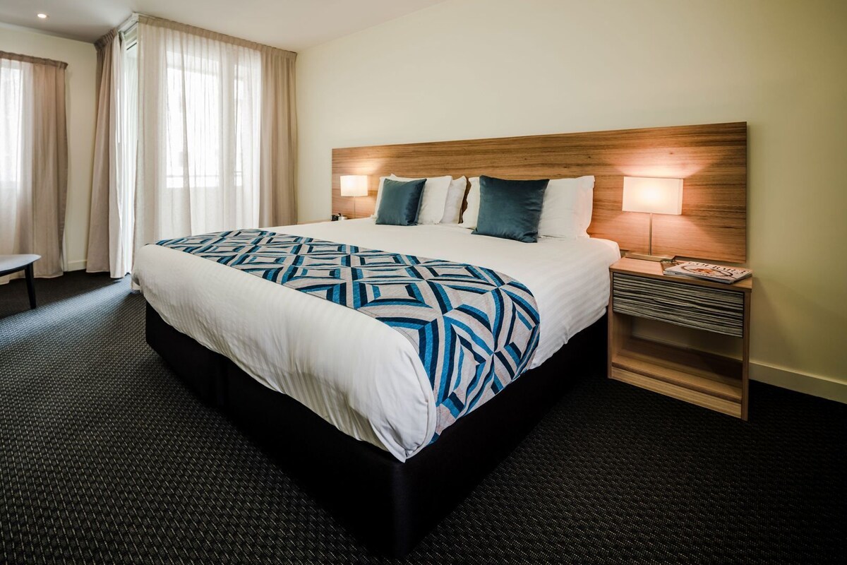 Ein Hotelzimmer mit einem großen Bett, einem Sessel, einem Nachttisch und einem Fenster mit Vorhängen.