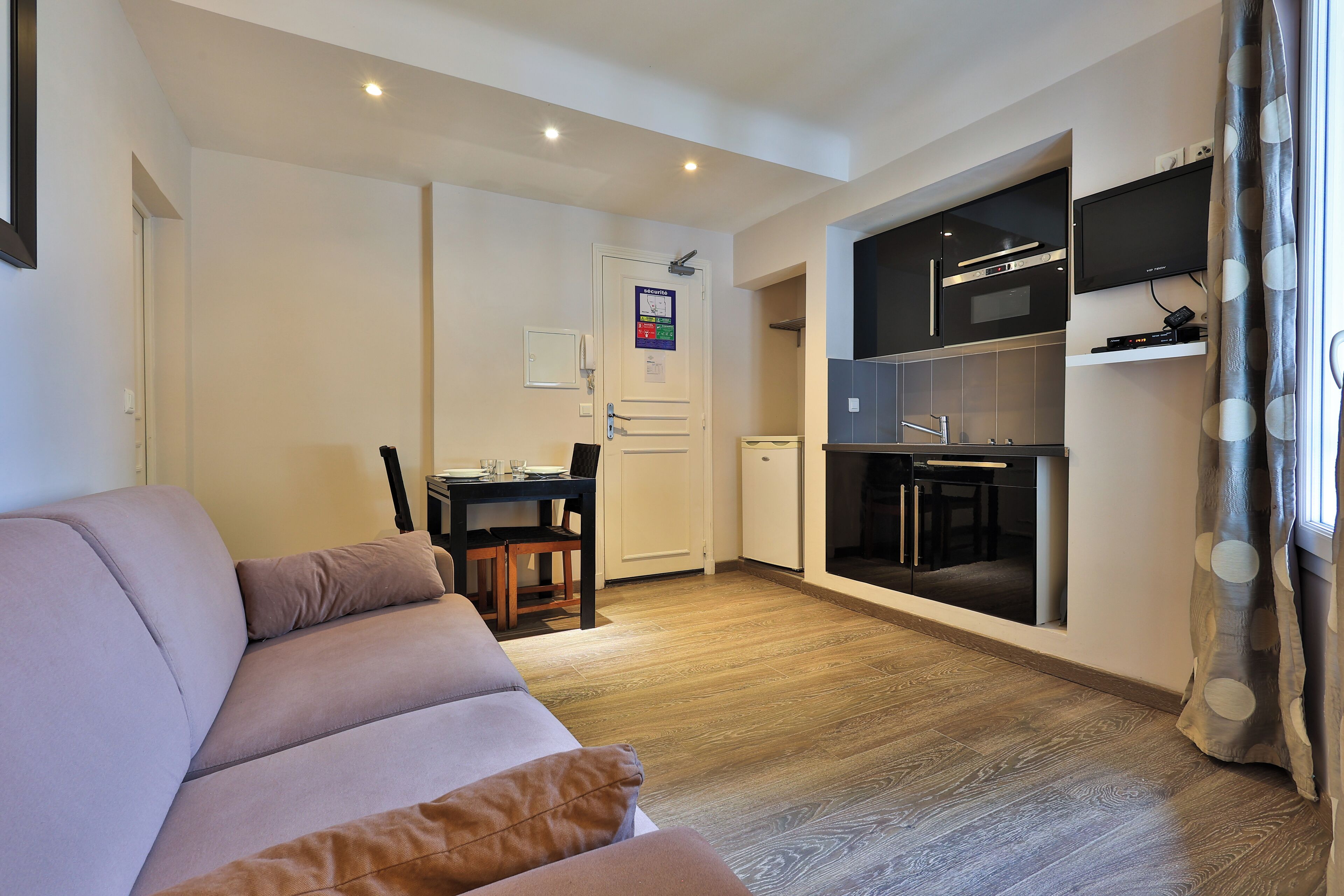 A modern living room with a sofa, a dining table with chairs, a kitchen area with a microwave and oven, and a TV mounted on the wall.