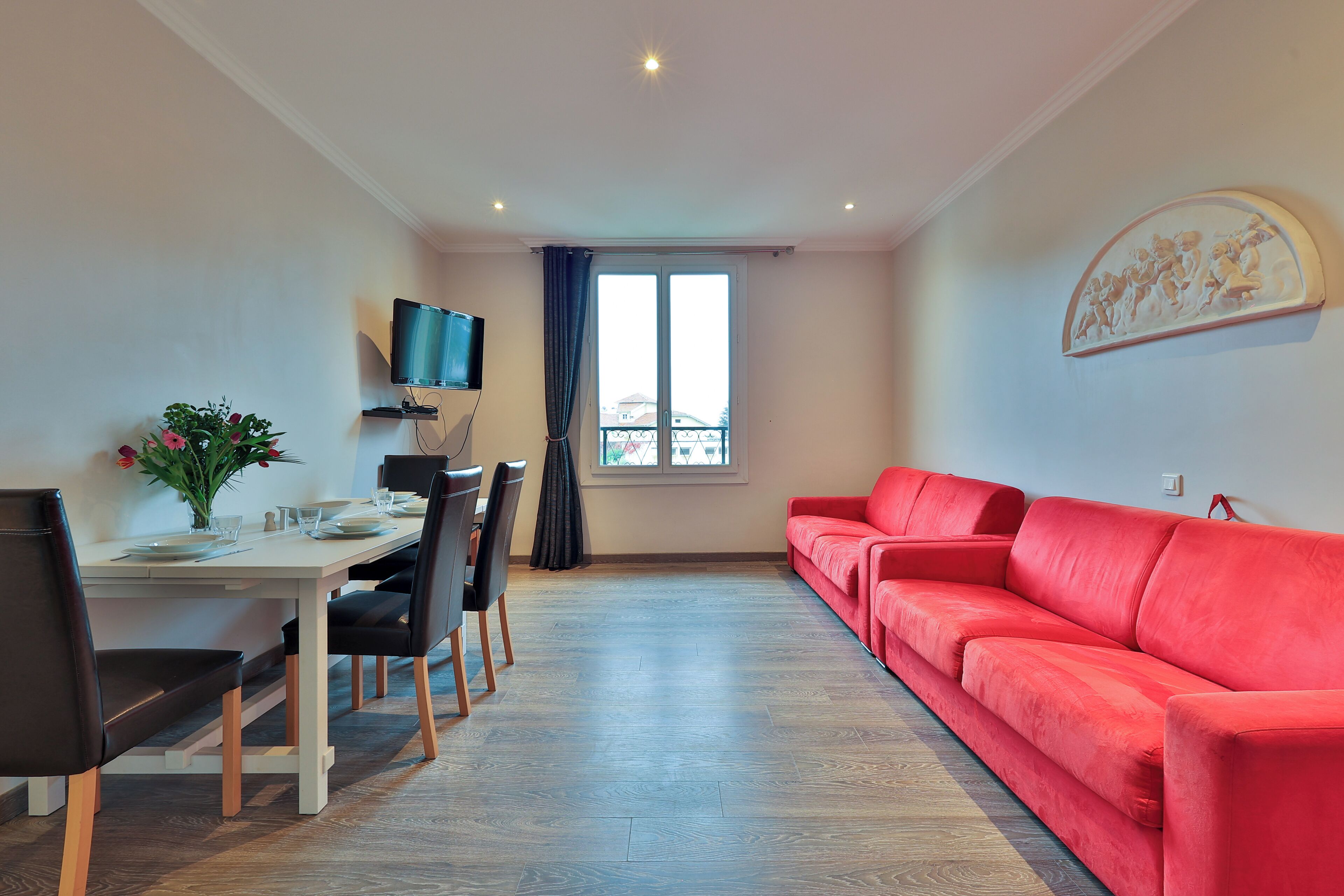 Una sala de estar moderna con un sofá rojo, una mesa de comedor con sillas, un televisor de pantalla plana montado en la pared y un ventanal con cortinas.