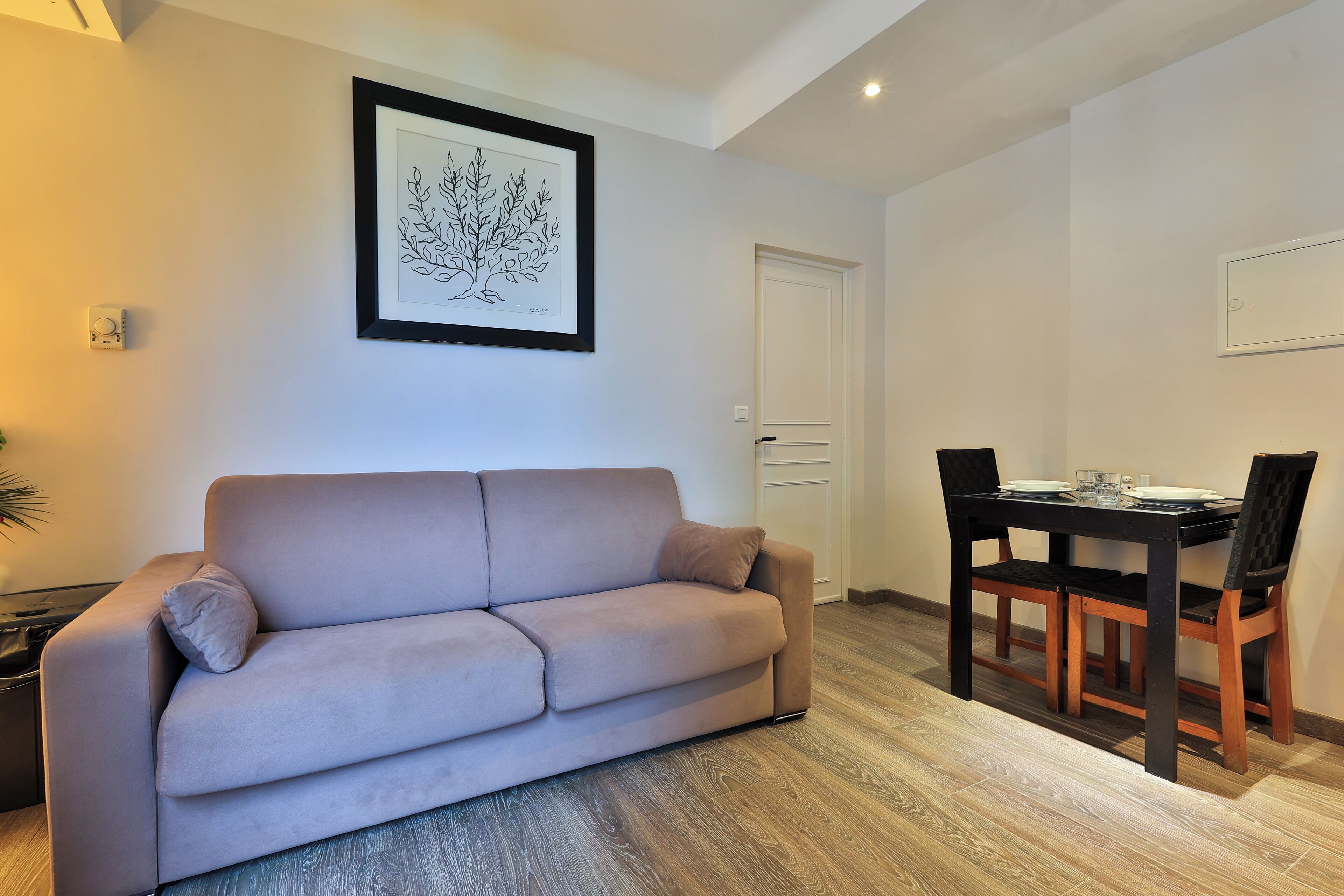 A living room with a sofa, a dining table with chairs, a framed artwork on the wall, and a door.
