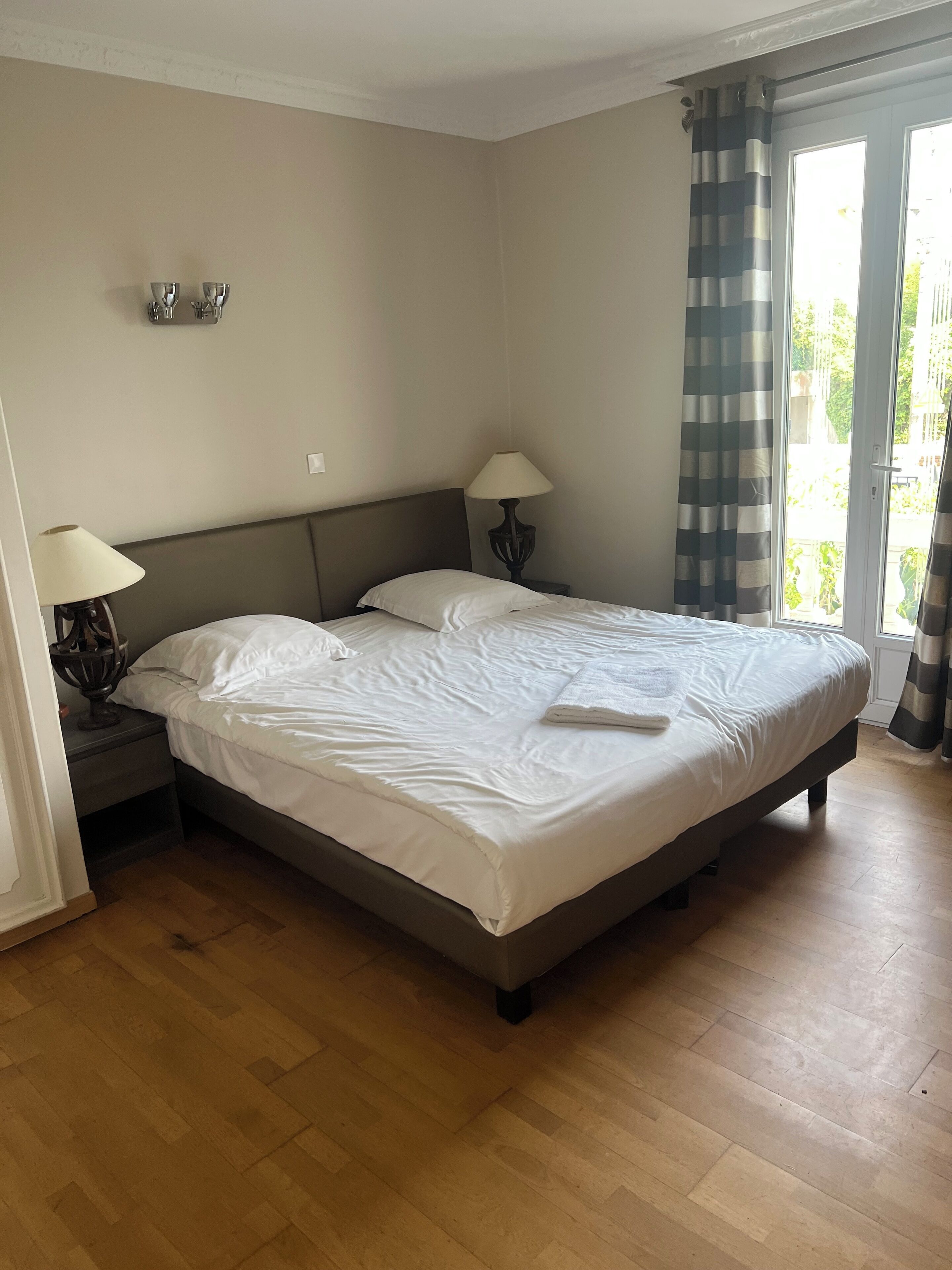 A bedroom with a large bed, two bedside tables with lamps, a window with curtains, and a view of greenery outside.