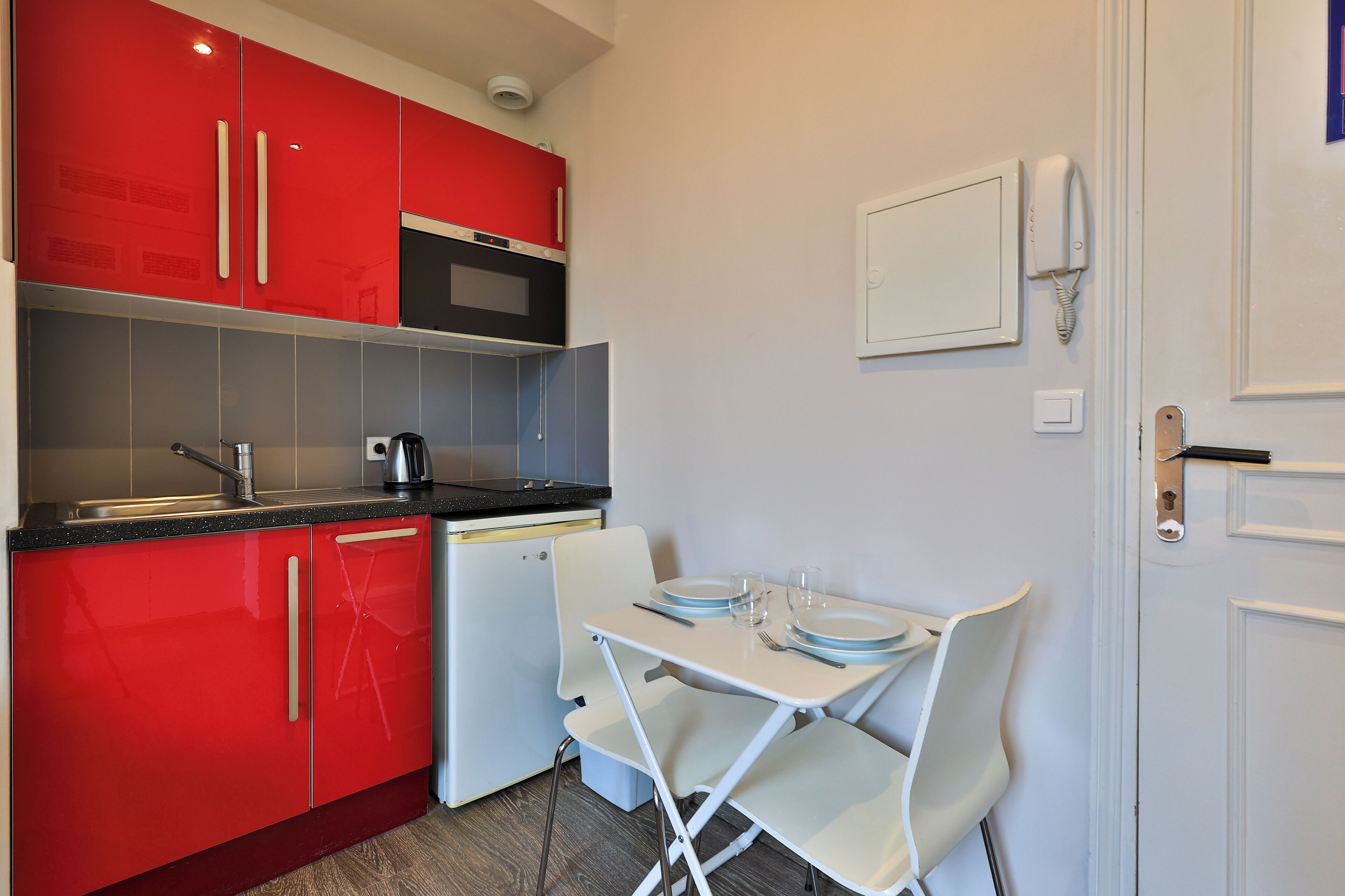 A compact kitchen with red cabinets, a microwave, a small refrigerator, and a foldable dining table with chairs.