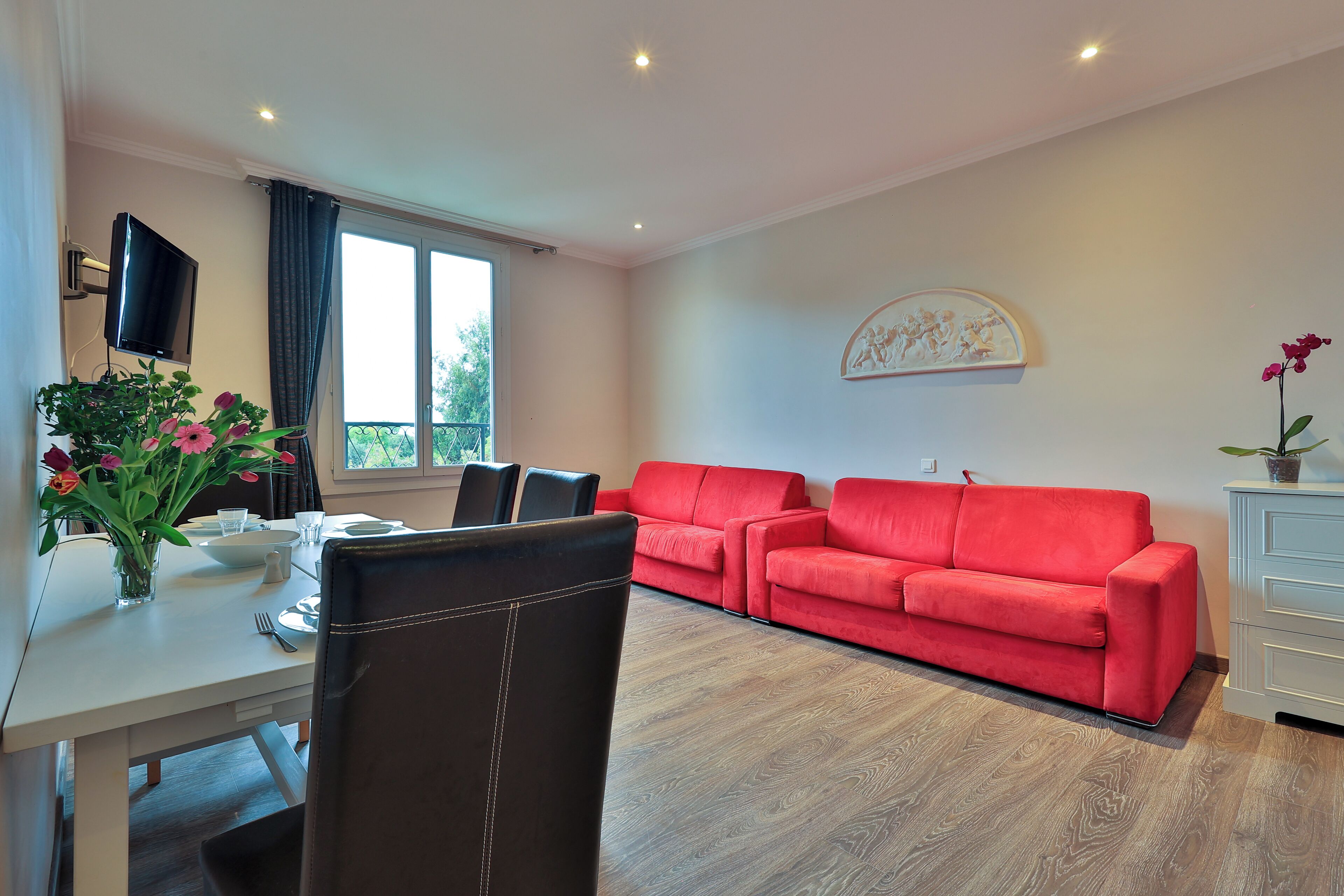 Una sala de estar moderna con un sofá rojo, una mesa de comedor con sillas, un televisor de pantalla plana montado en la pared y una ventana con cortinas.