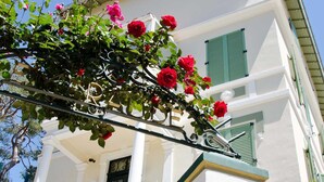 Un edificio blanco con contraventanas verdes, una valla de metal y flores rojas.