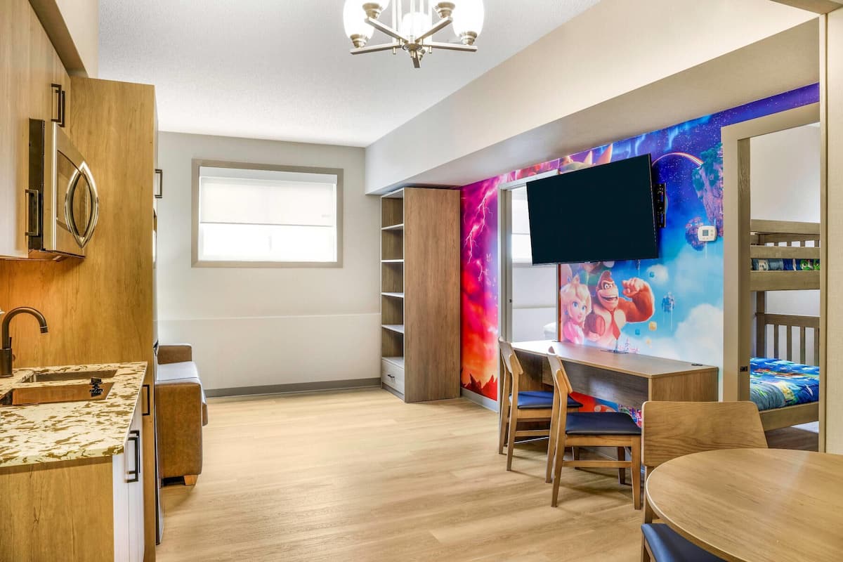 A modern children's room with a wooden kitchenette, a TV mounted on a vibrant mural, a bunk bed, and a desk with chairs.