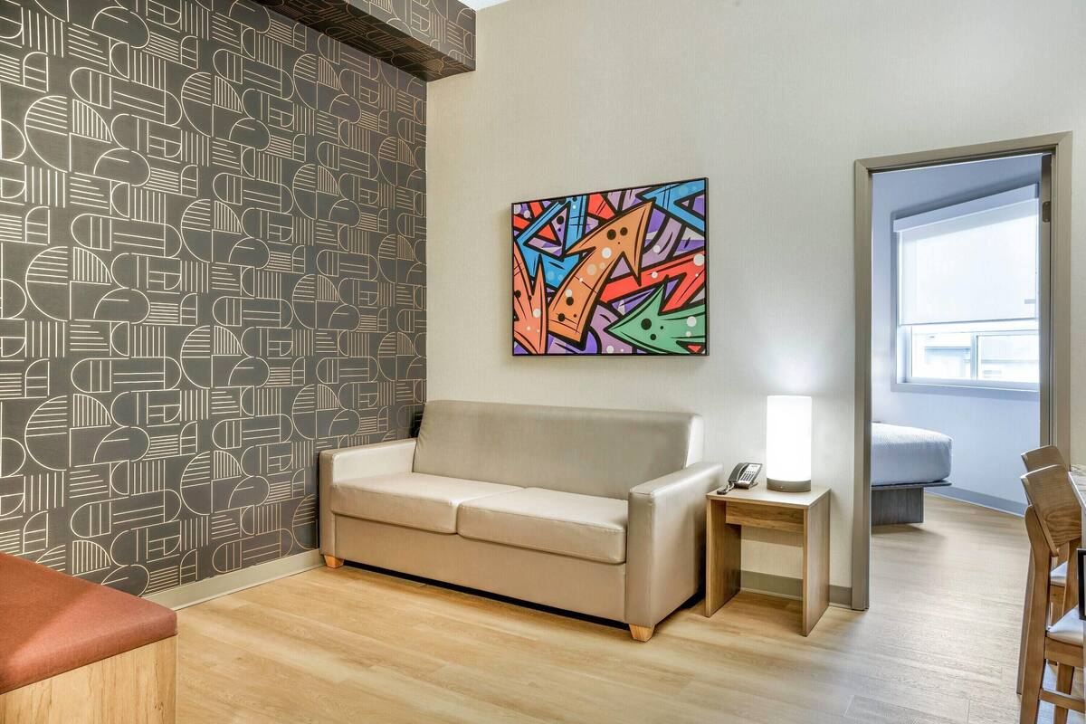 A modern room with a patterned wall, a beige sofa, a small bedside table with a lamp, and a view of a balcony.