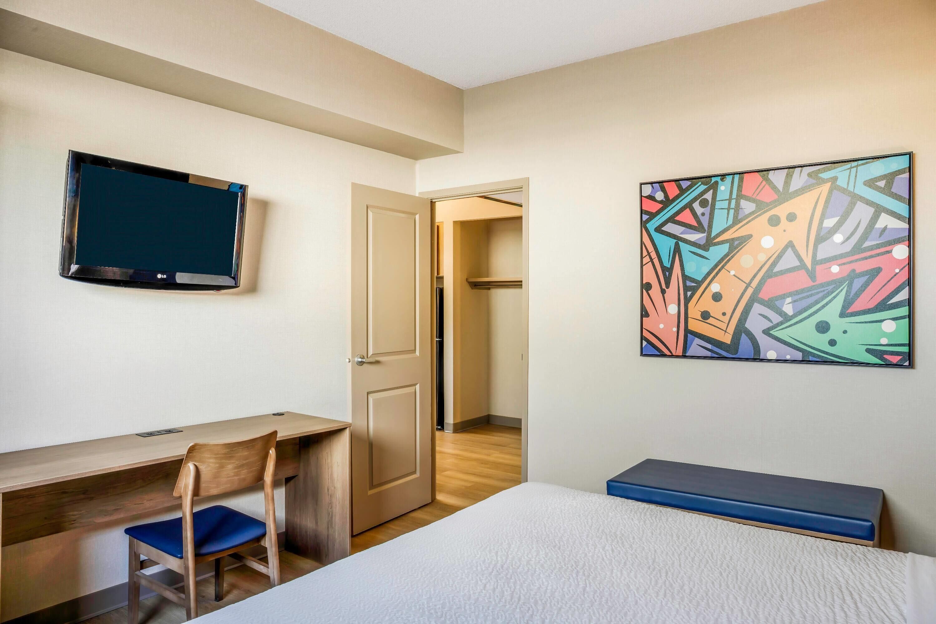 Una habitación de hotel con cama, escritorio con silla, televisor montado en la pared y un cuadro abstracto de colores.