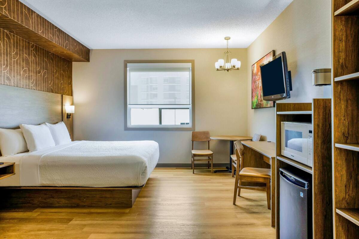 A hotel room with a bed, a desk, a chair, a TV, a microwave, and a refrigerator.