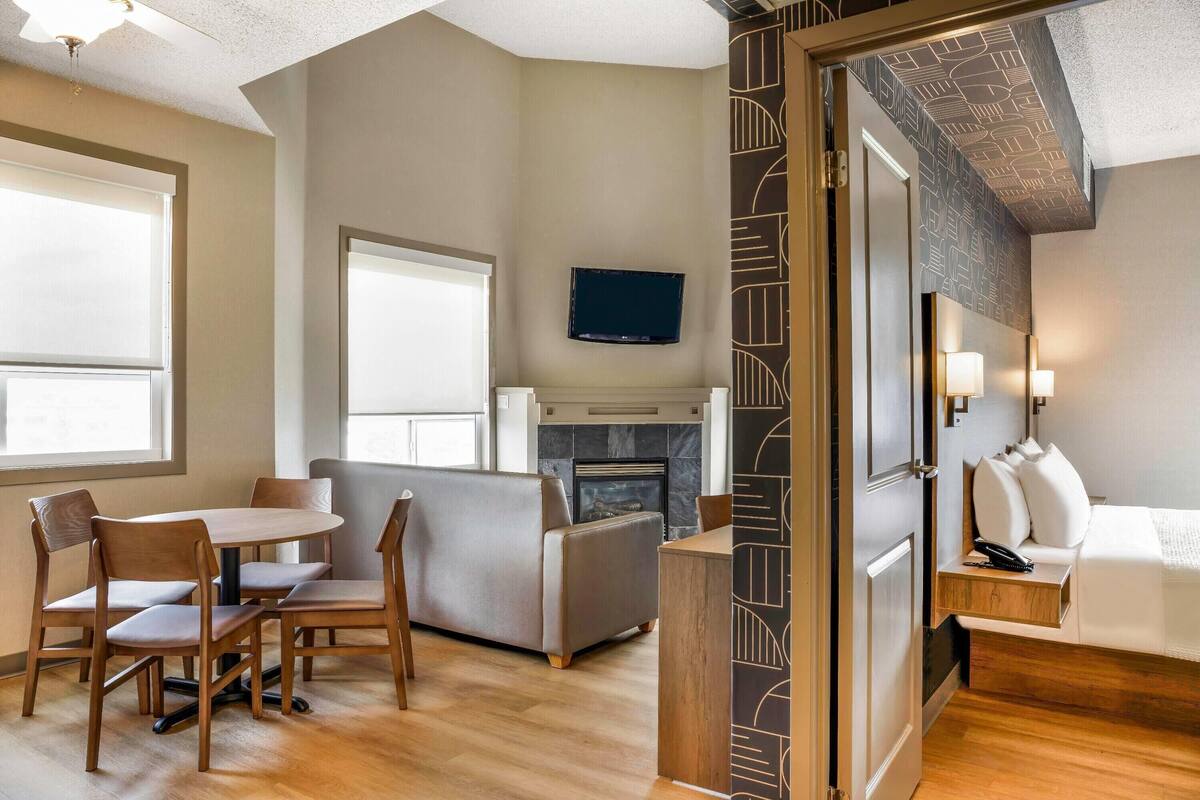 A hotel room with a dining area, a sofa, a TV mounted on the wall, and a fireplace.