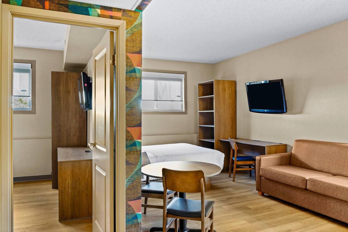 A compact room with a bed, a TV mounted on the wall, a wooden desk, a brown sofa, and a small table.