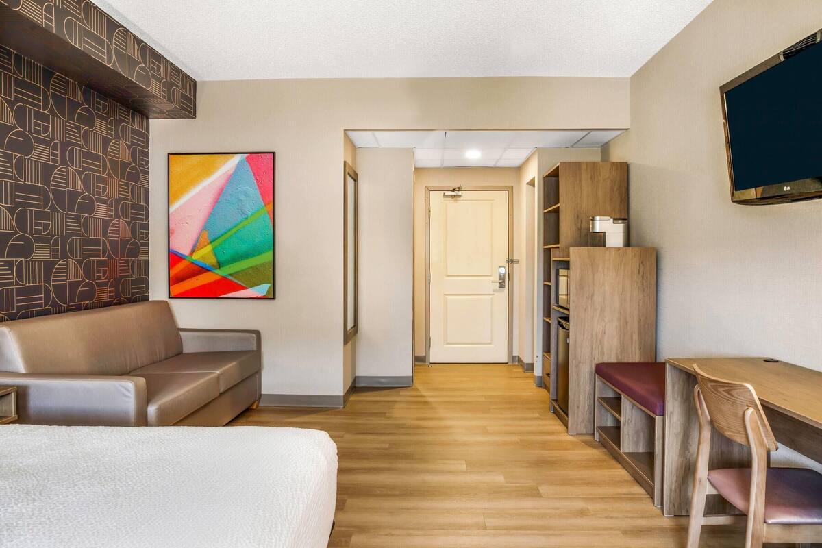 A hotel room with a bed, a sofa, a desk with a chair, a TV, and a colorful abstract painting on the wall.