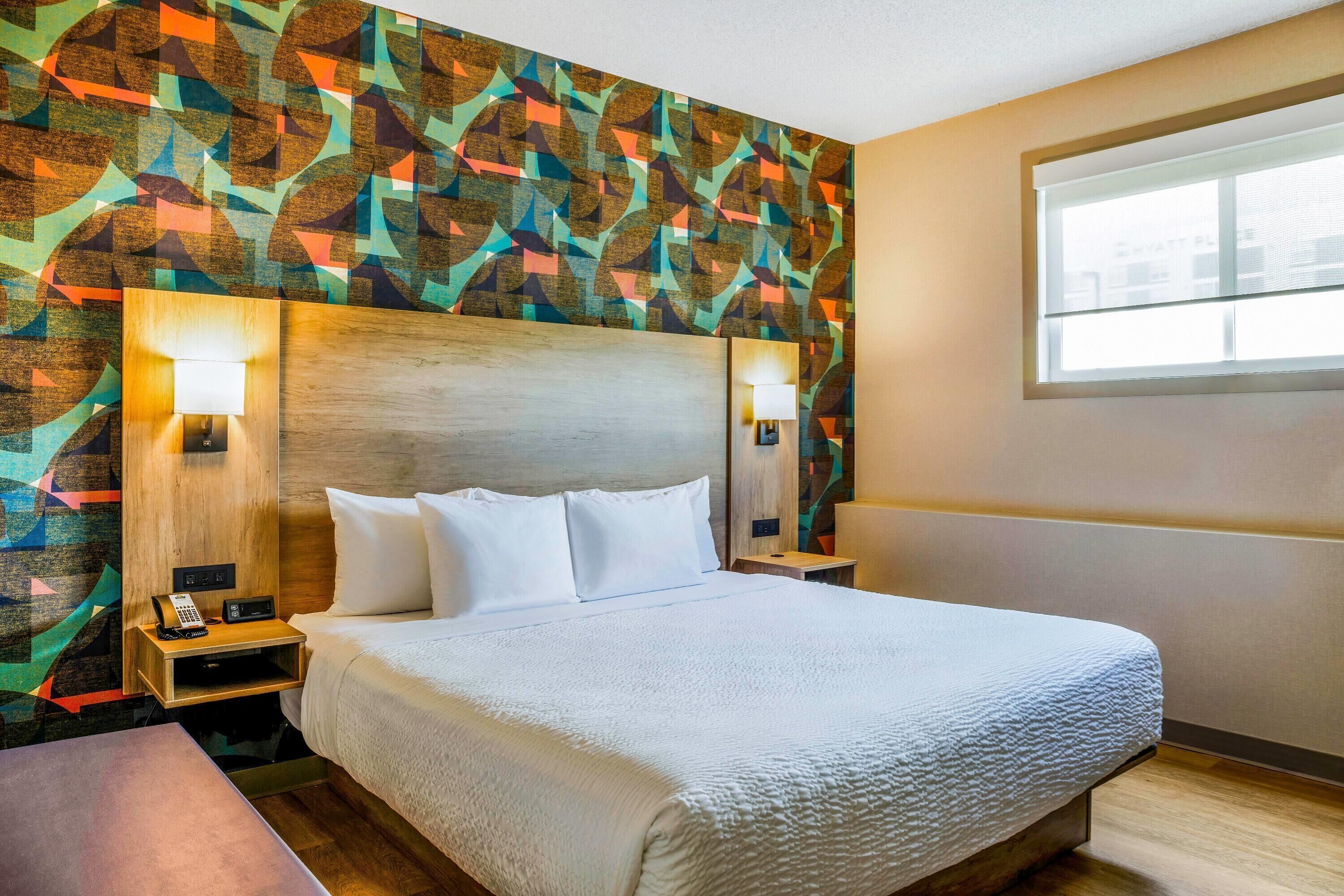 A hotel room with a large bed, two bedside tables, a nightstand, a patterned wall, and a window.