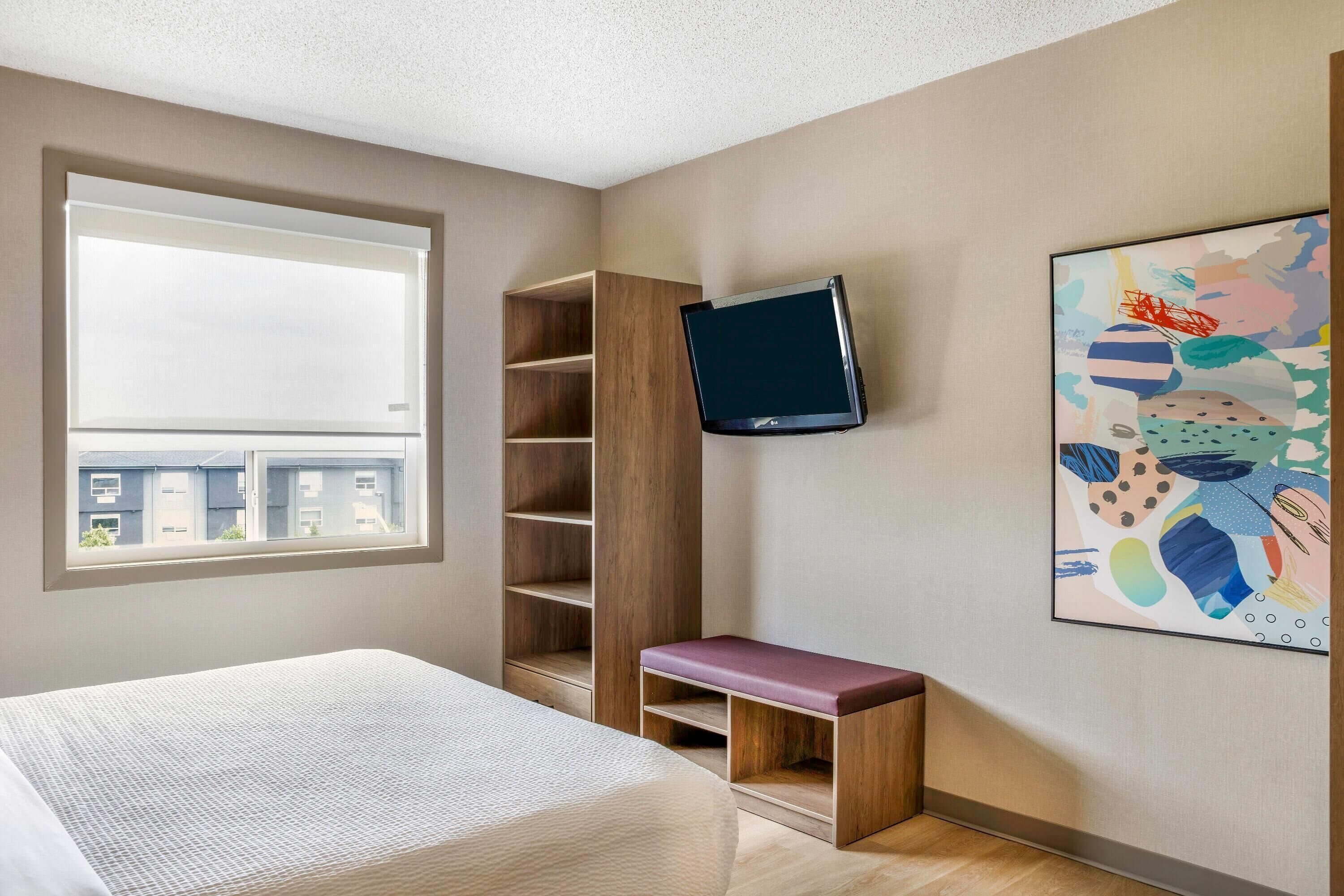 A bedroom with a bed, a TV mounted on the wall, a bench, and a window with a view of buildings.