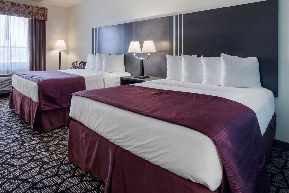 A hotel room with two beds, a large dark headboard, two bedside lamps, and a patterned carpet.