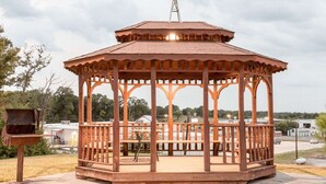 Um gazebo de madeira com um moinho de vento no topo, cercado por árvores e um corpo d'água.