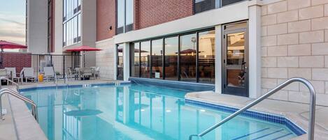 An outdoor swimming pool with a metal handrail, surrounded by a brick building with large windows and a covered seating area with umbrellas.