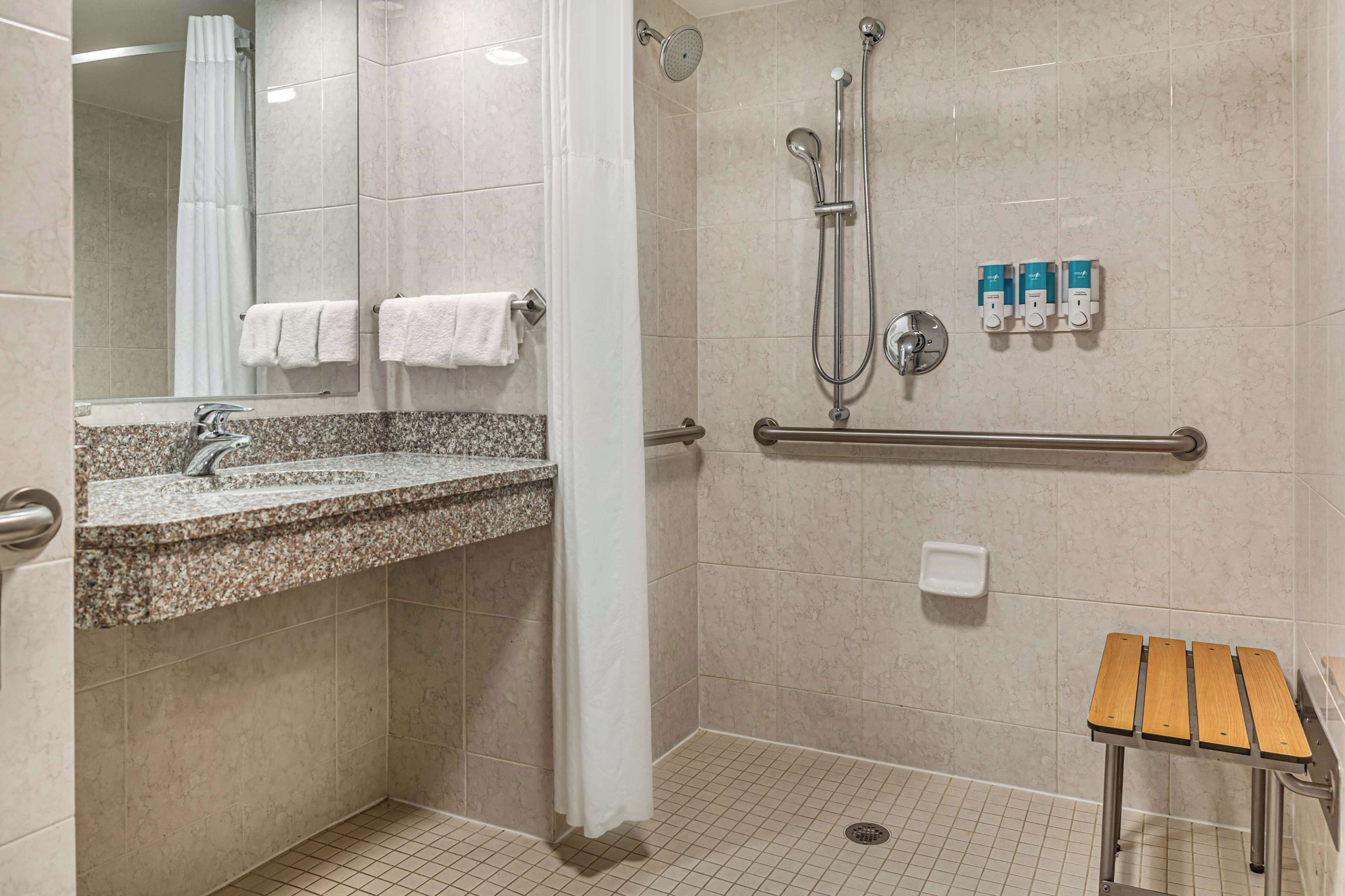 Un baño moderno con lavamanos de granito, ducha con cabezal de ducha manual, barra de apoyo y banco de madera.