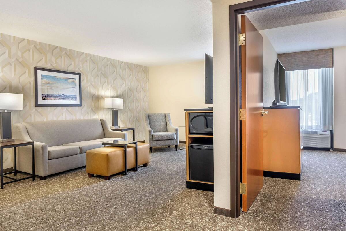 Ein Hotelzimmer mit Sofa, Hocker, Ohrensessel und einem Fernseher auf einem Standfuß.